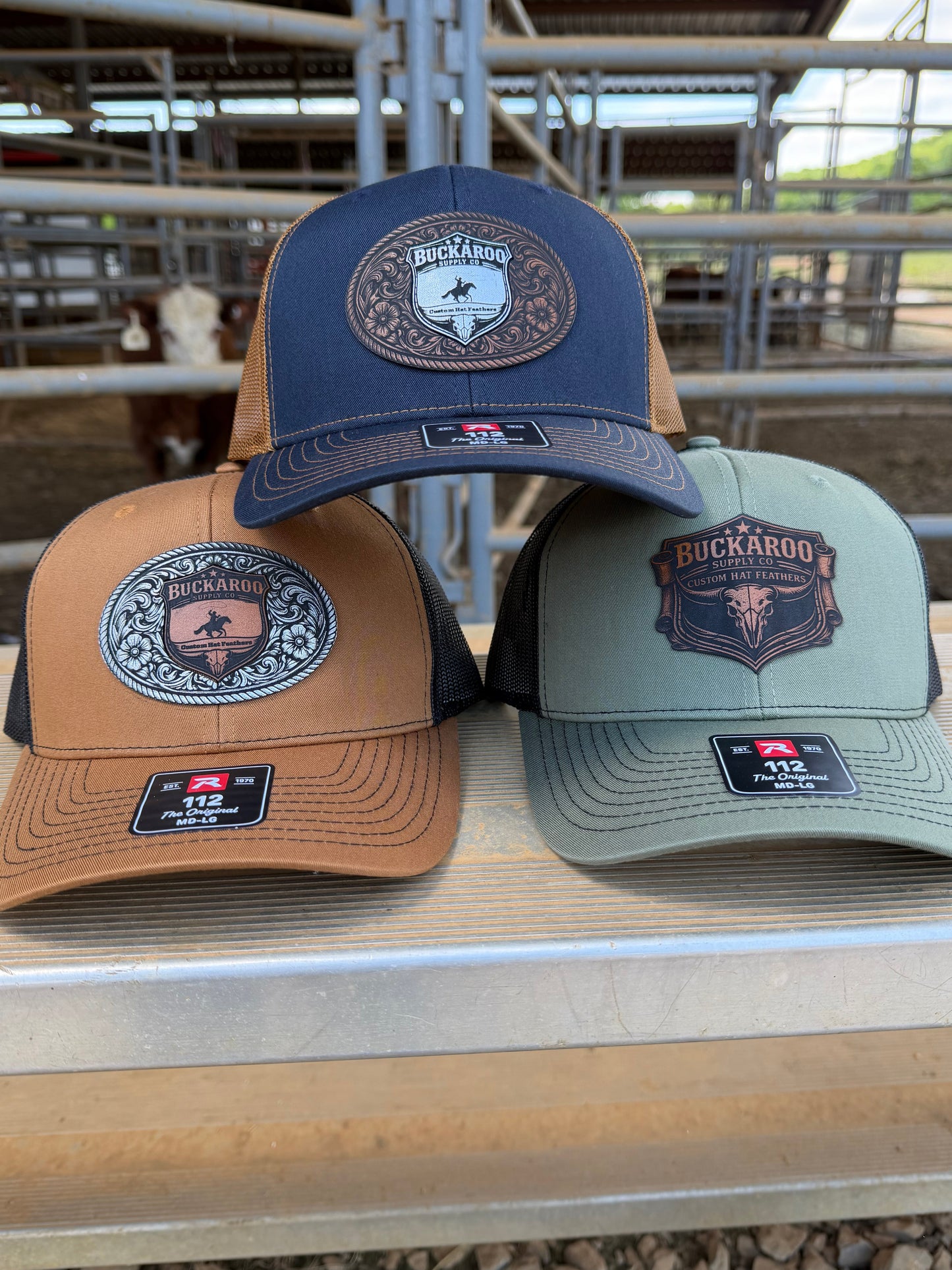 Three caps in blue, green, and brown with logos on a wooden surface. Loden (sage) Green and black, Richardson 112 cap with Buckaroo Supply Co logo.