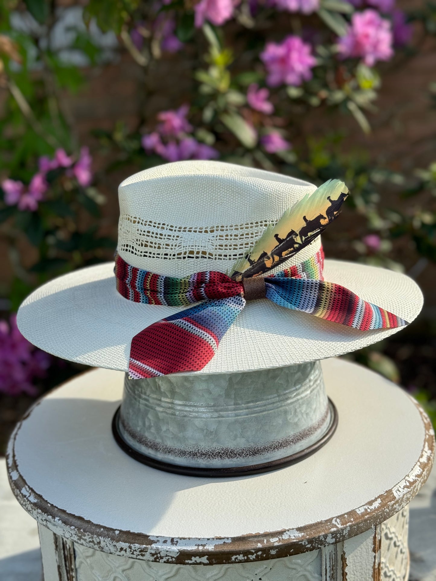 White hat with a colorful ribbon and Buckaroo Supply Co. hat feather on a rustic wooden stand with blurred floral background