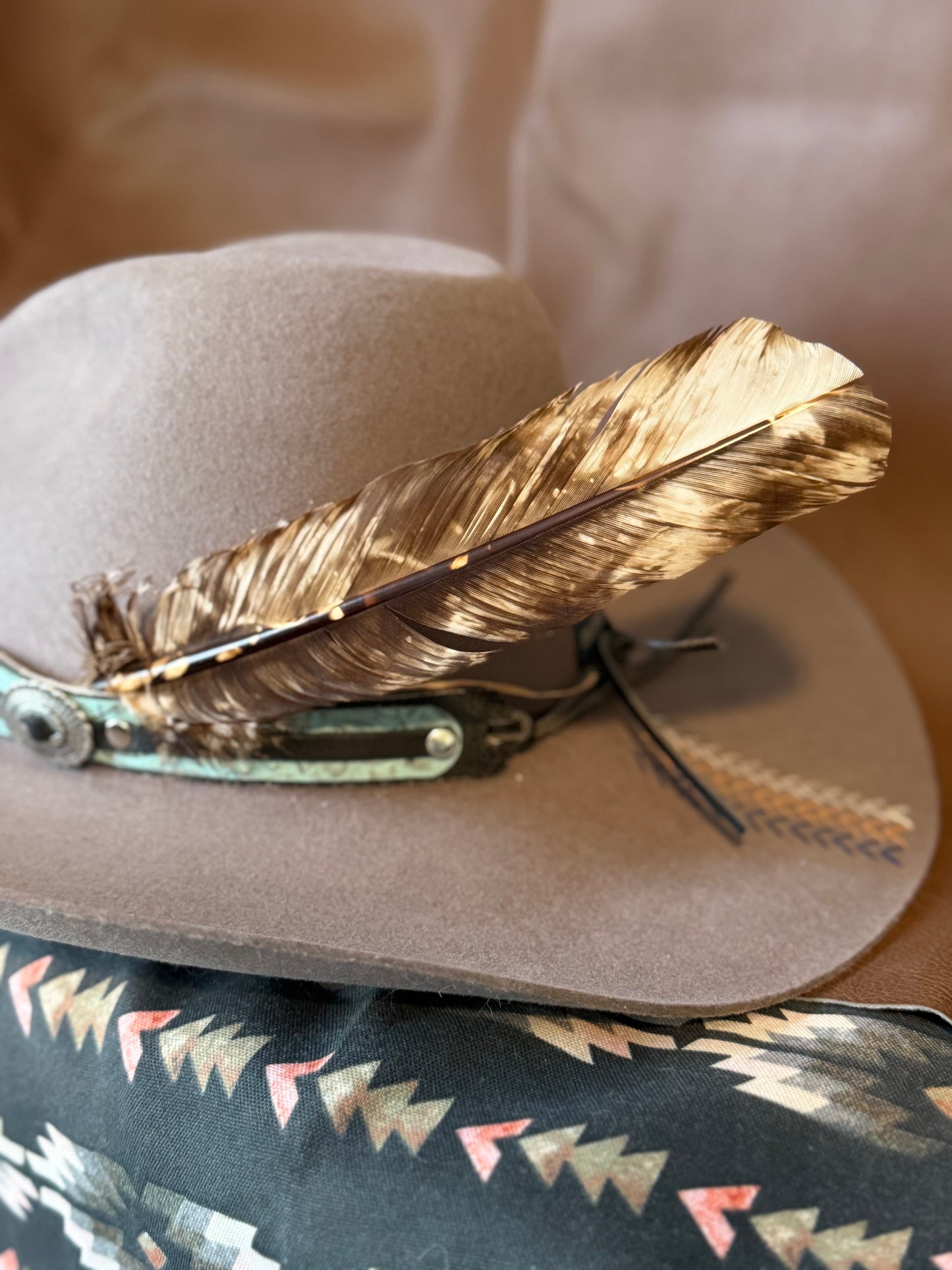 A light brown cowboy hat with a large brown and gold feather and turquoise concho detail on the side, resting on a patterned black fabric.