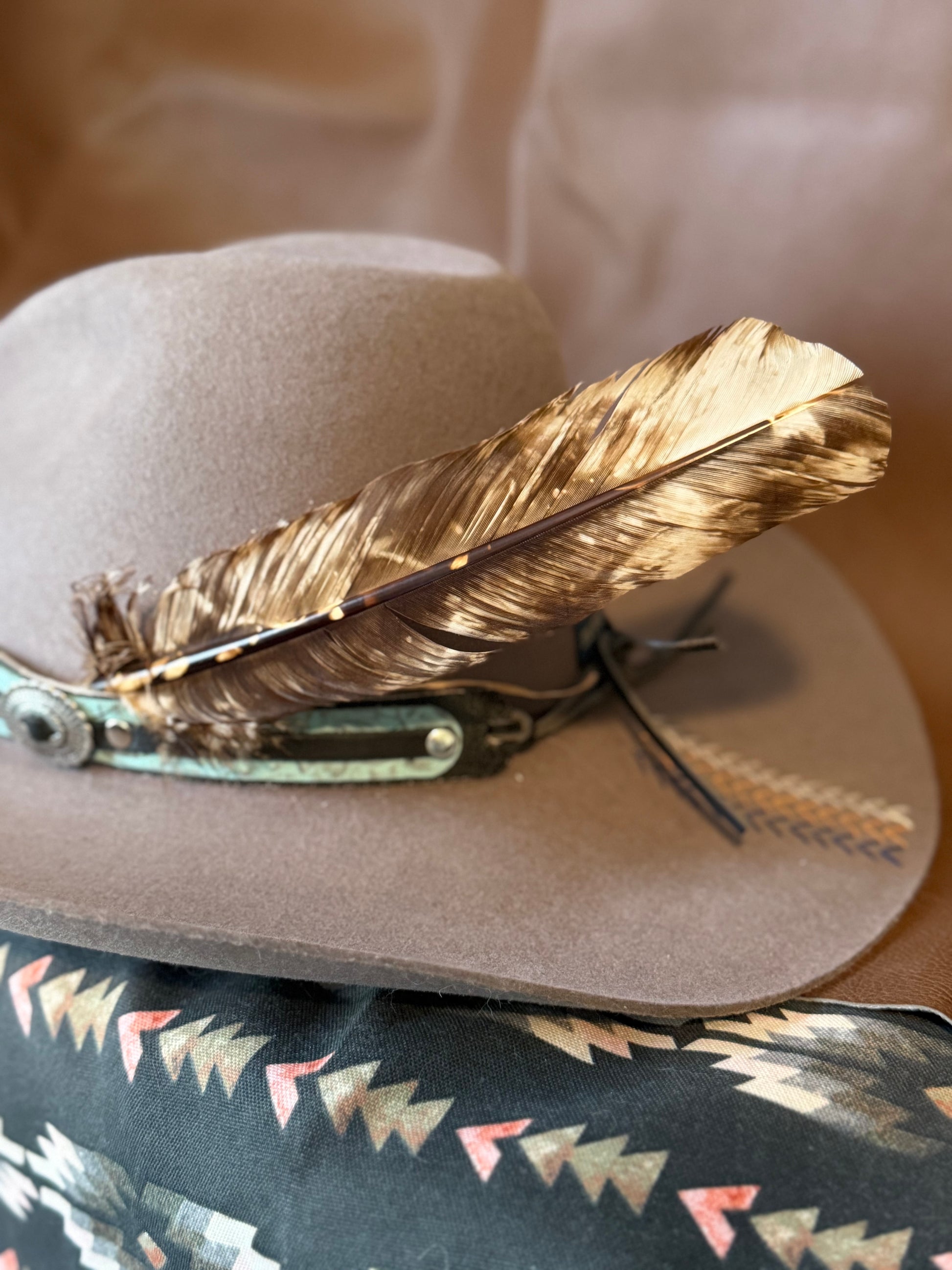 A light brown cowboy hat with a large brown and gold feather and turquoise concho detail on the side, resting on a patterned black fabric.