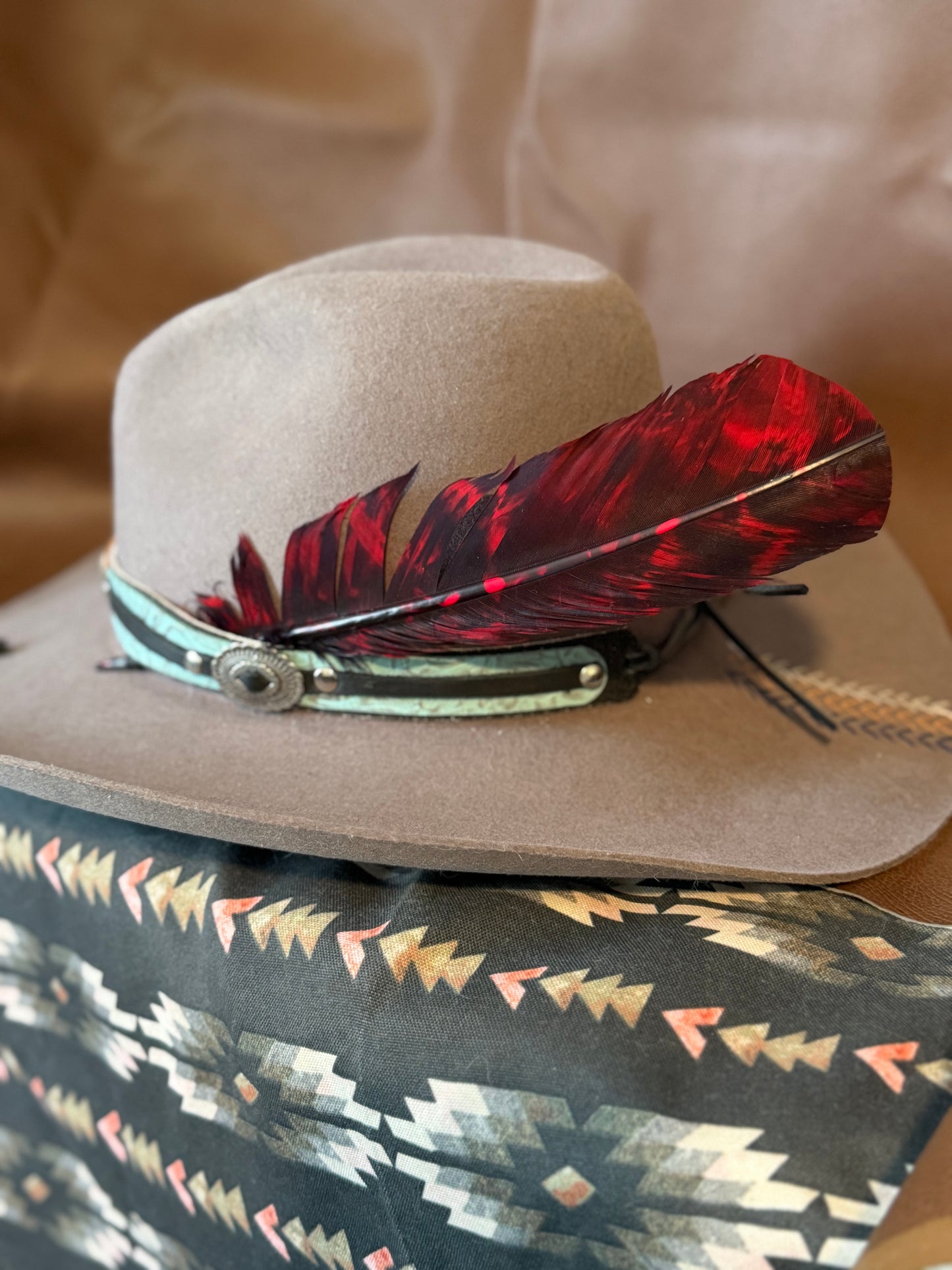 A light brown felt cowboy hat with a turquoise and black band, decorated with a large red and black feather, resting on a patterned textile.