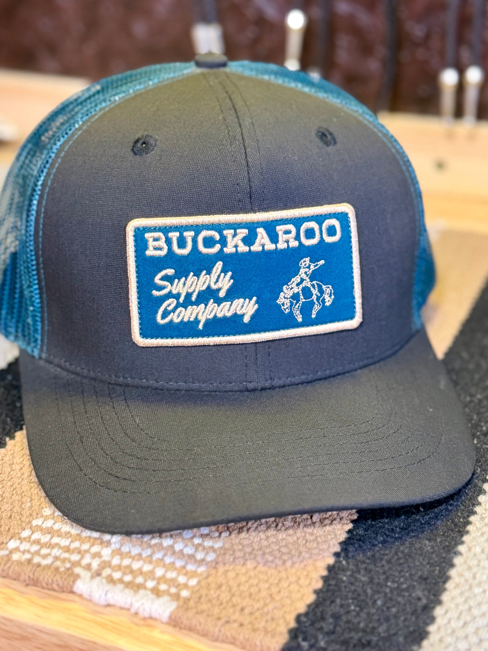 A trucker-style baseball cap with a dark charcoal gray front panel and brim, teal mesh back, and a rectangular embroidered patch reading 'BUCKAROO Supply Company' with a white cowboy graphic.