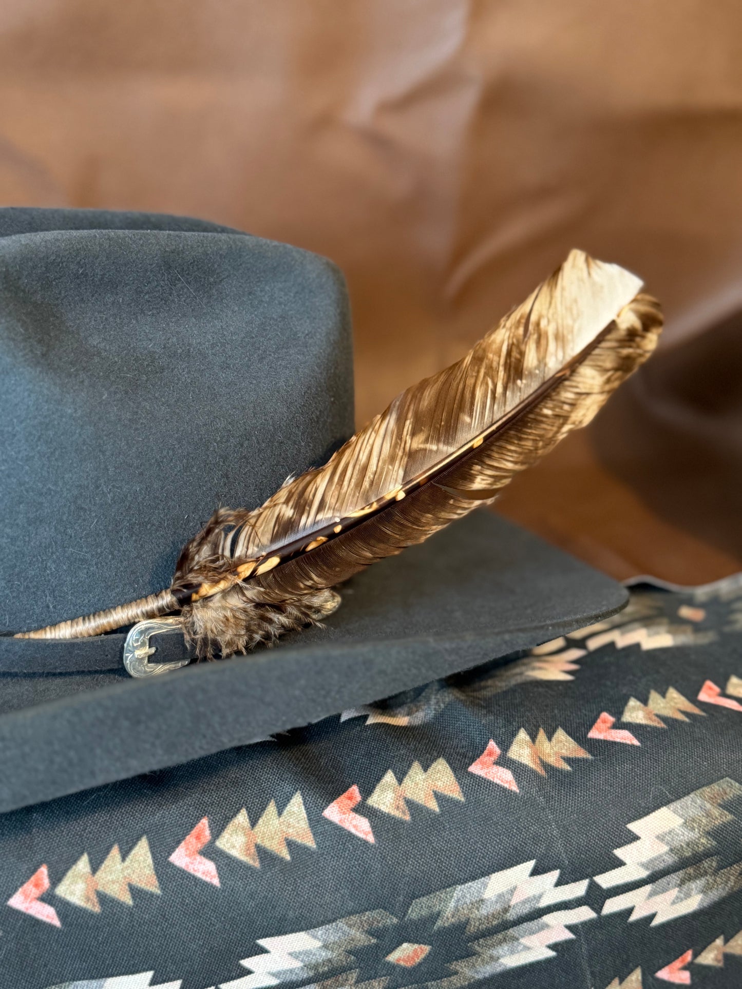 A close-up of a dark gray cowboy hat with a brown and gold feather resting on its brim, set against a brown leather-like background with a patterned textile below.