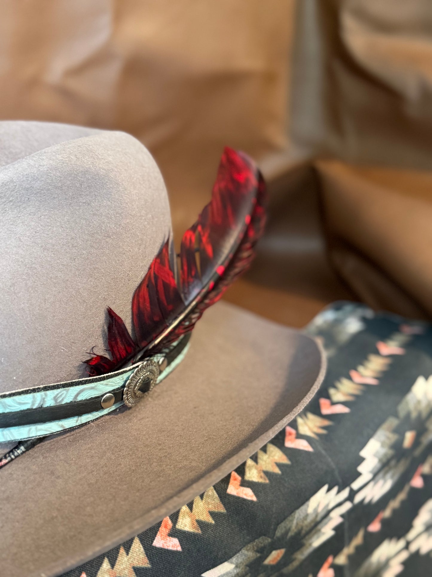 A close-up of a light gray felt cowboy hat with a turquoise and black patterned band and a red feather accent, resting on a patterned surface.