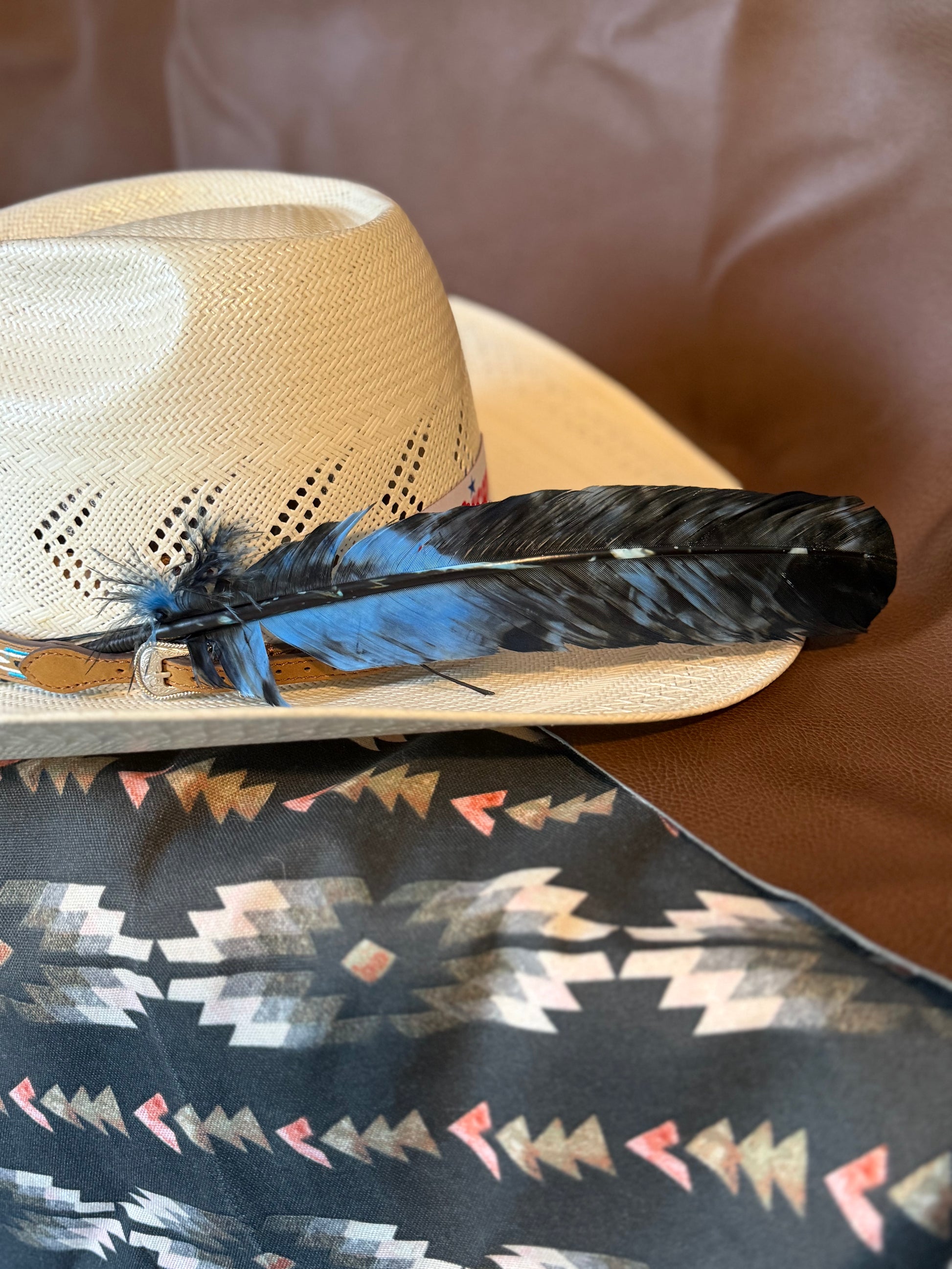 A light beige straw cowboy hat with a long black and blue feather resting across the brim, placed on top of a dark patterned fabric against a brown leather background.