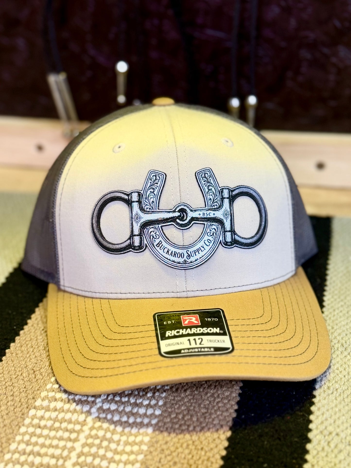 Front view of Mink Beige Charcoal and Amber Gold Richardson 112 trucker hat with Buckaroo snaffle bit and horseshoe artwork