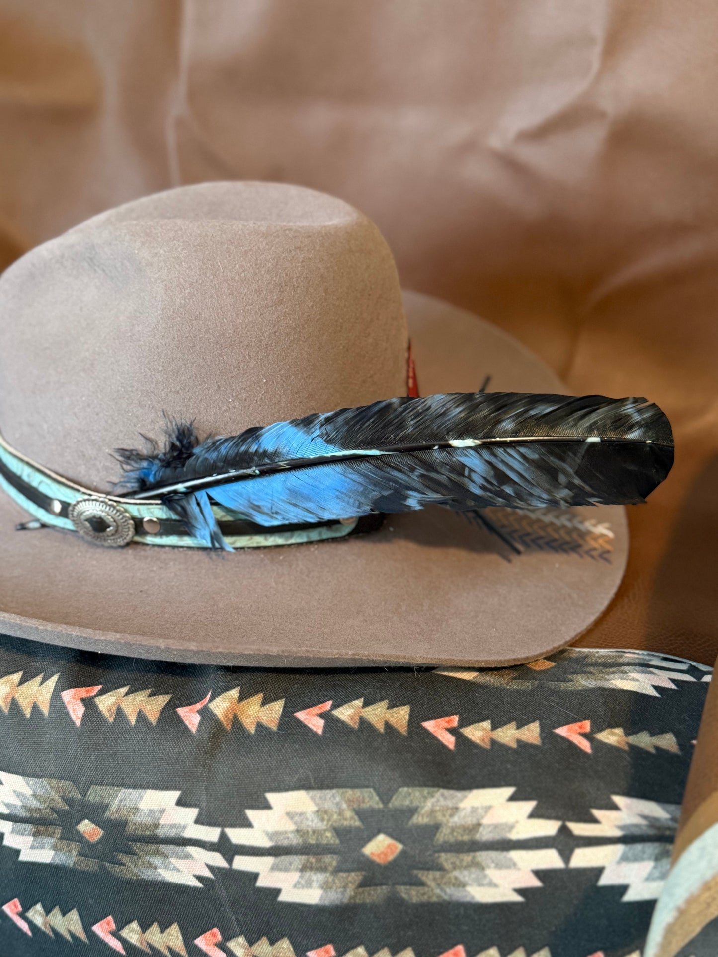 A light brown cowboy hat with a turquoise and black feather band, resting on a patterned southwestern-style pillow.
