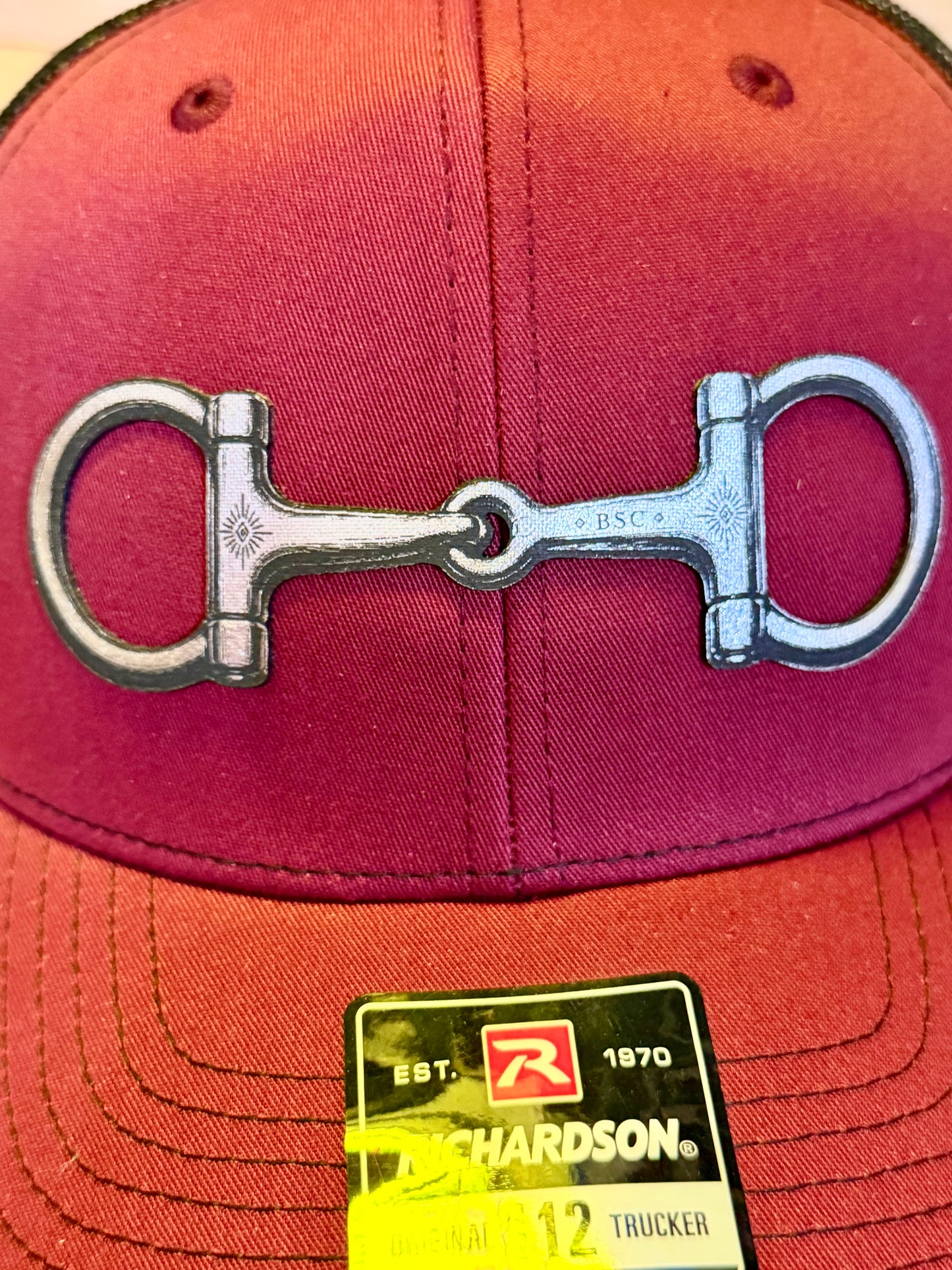 A close-up of a red baseball cap featuring a large silver horse bit as the main design element, with a yellow and black Richardson brand tag attached at the brim.