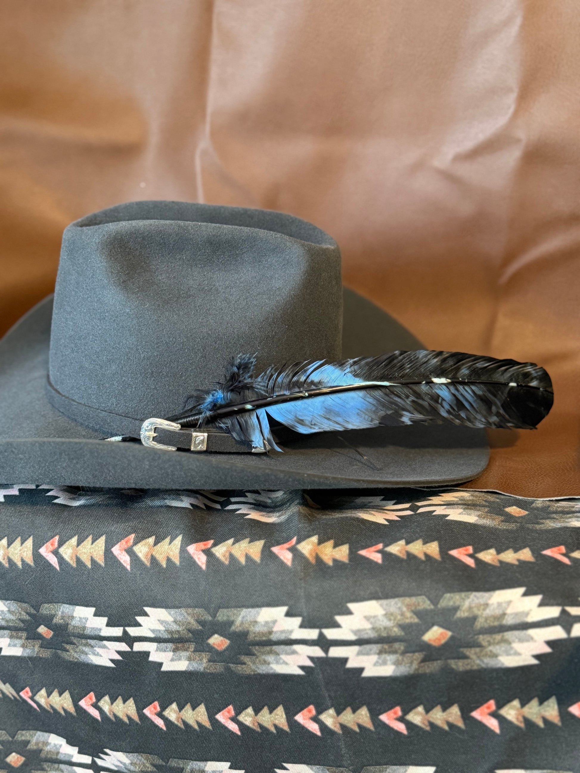 A dark gray felt cowboy hat with a black and blue feather accent, displayed on top of a patterned textile with a brown fabric backdrop.