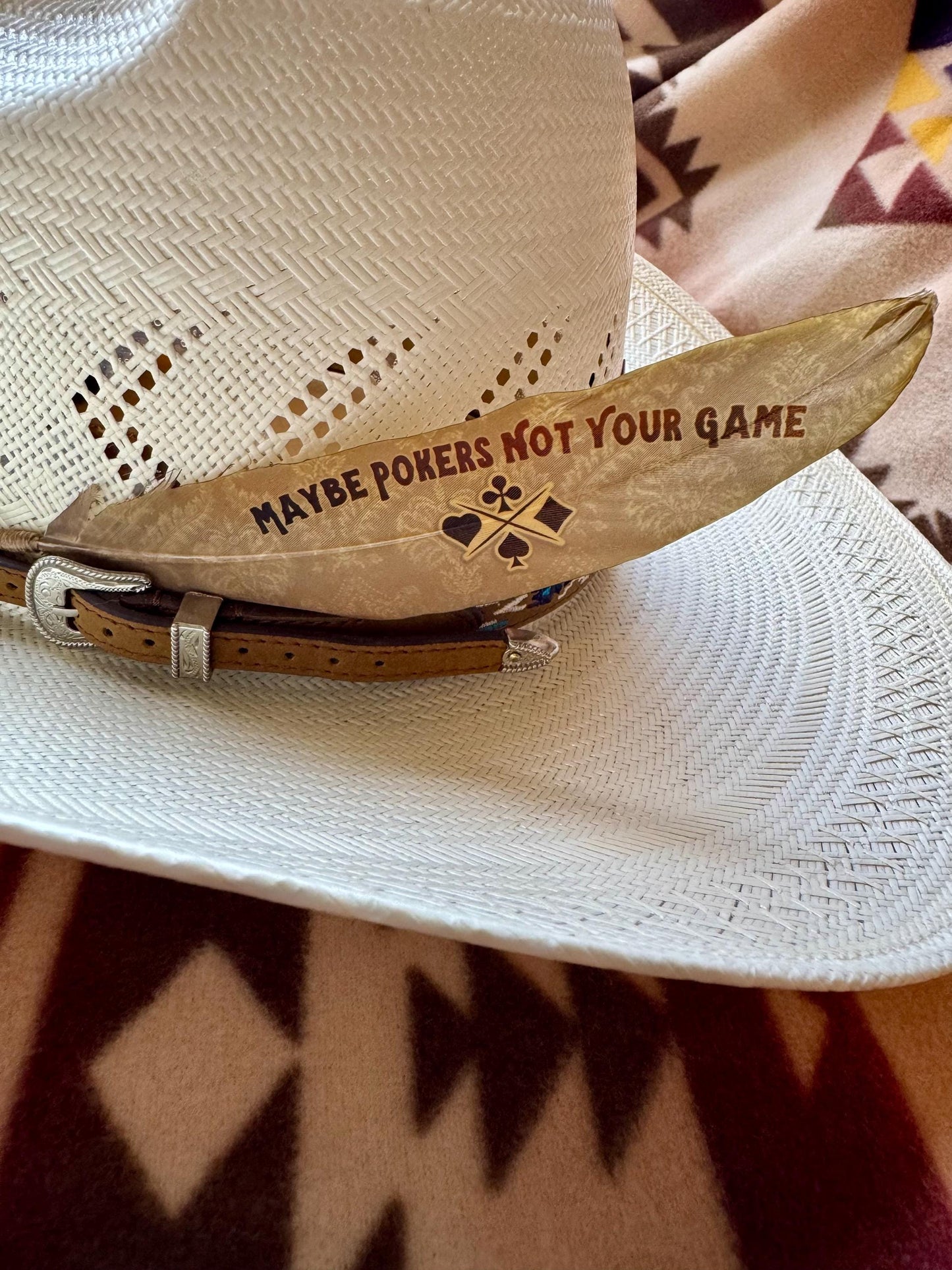 Cowboy hat with a wooden belt buckle that says 'Maybe Pokers Not Your Game' on a patterned fabric background.