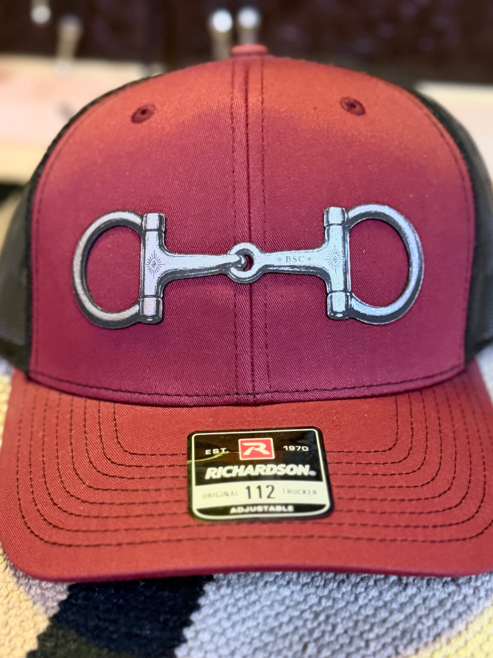 Close-up of silver-tone snaffle bit emblem on cardinal Richardson 112 trucker hat