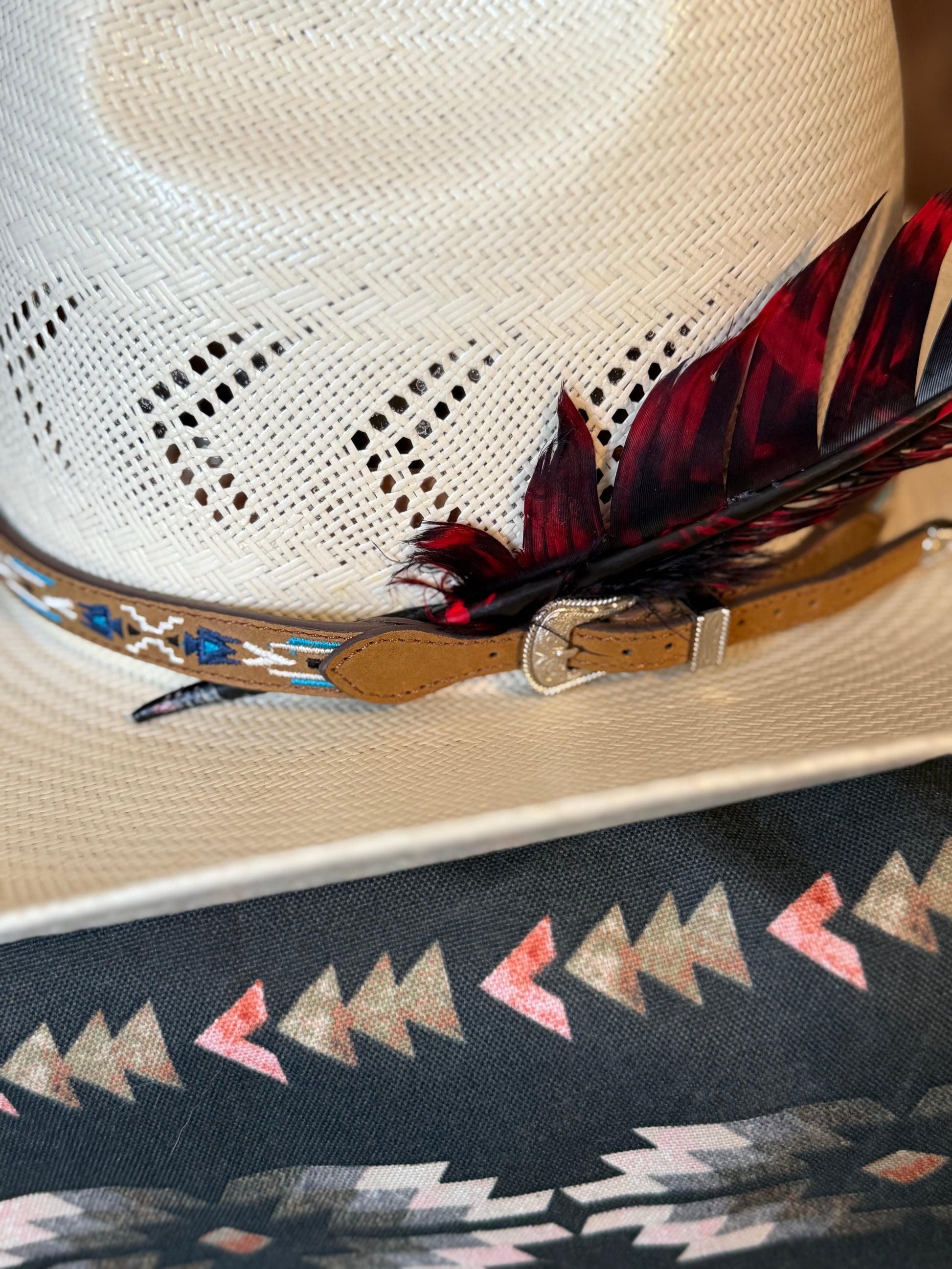 A close-up of a cream woven cowboy hat with a tan leather band, silver buckle, and dark red and black feather, resting on a patterned textile.