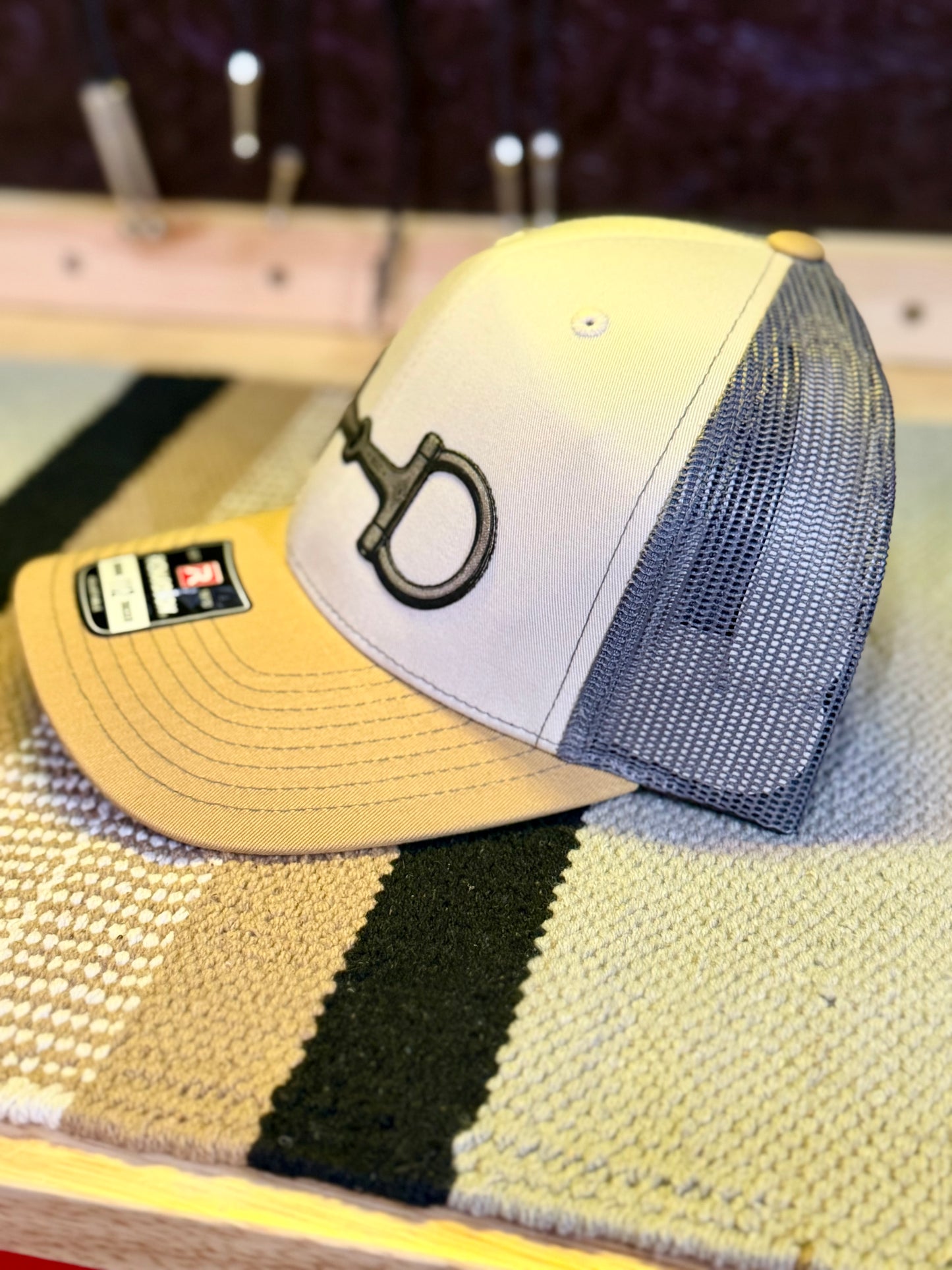 A trucker-style baseball cap with a light beige front panel, a tan brim, and a gray mesh back, featuring a silver snaffle bit patch