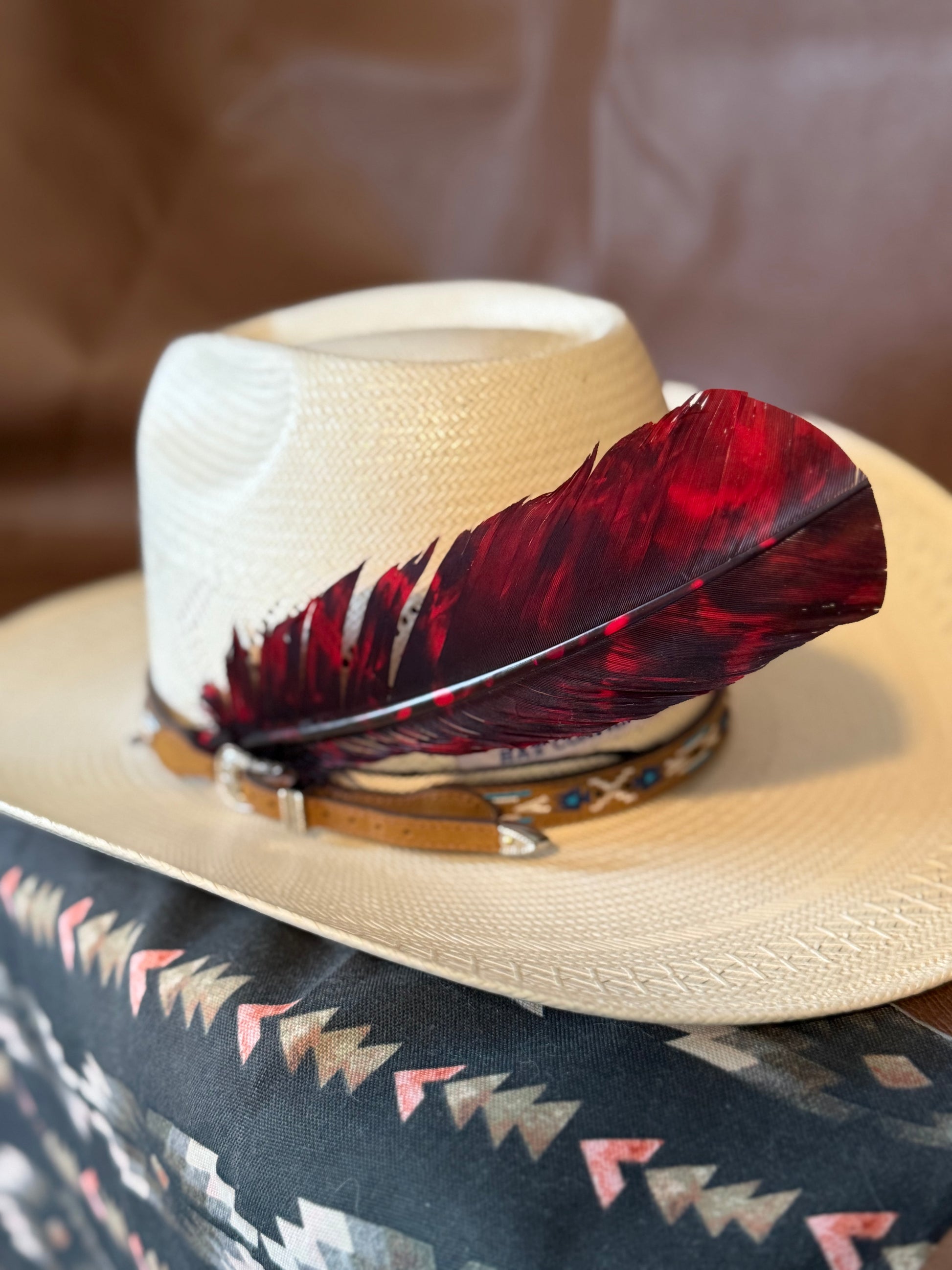 A light beige straw cowboy hat with a large dark red and black feather and a tan leather band, resting on a patterned textile.