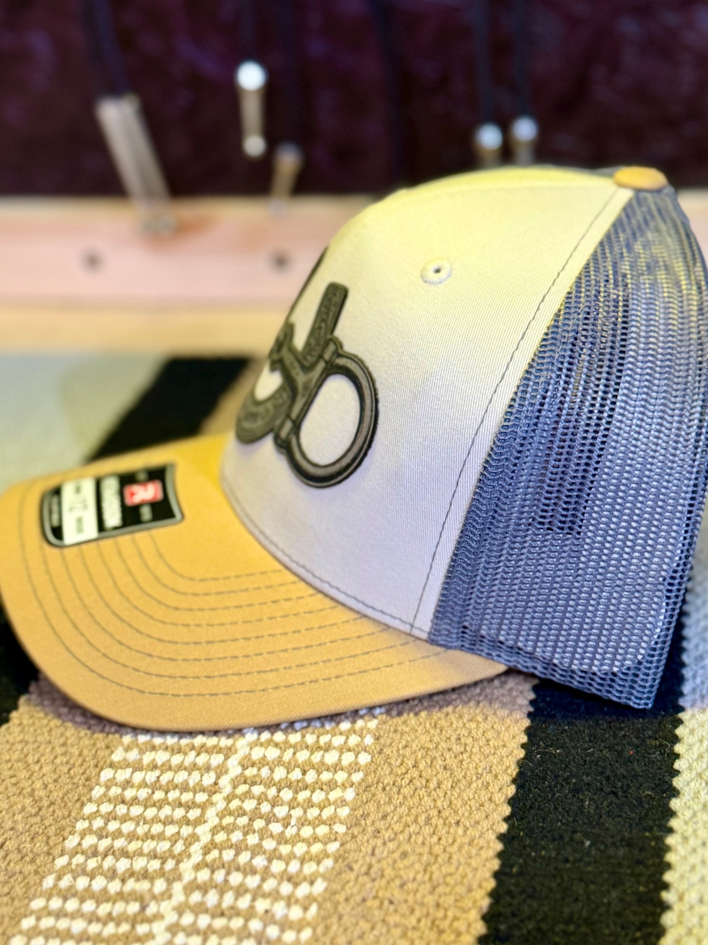 A trucker-style baseball cap with a white front panel, blue mesh back, and tan visor, featuring a silver snaffle bit and horseshoe silver leather patch on the front.