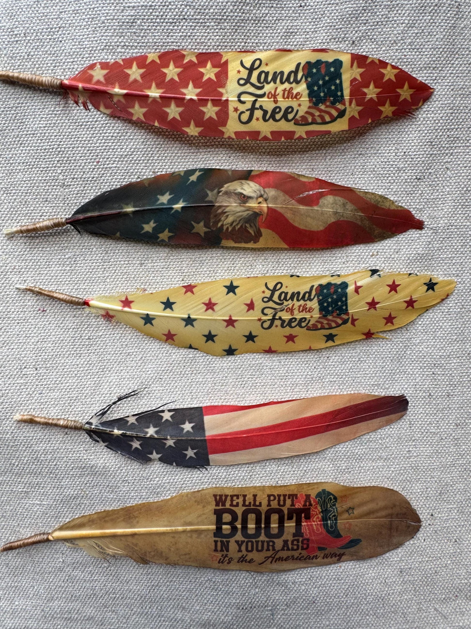Decorative feathers with American flag and text designs on a textured surface, Buckaroo Supply Co. Western hat feather