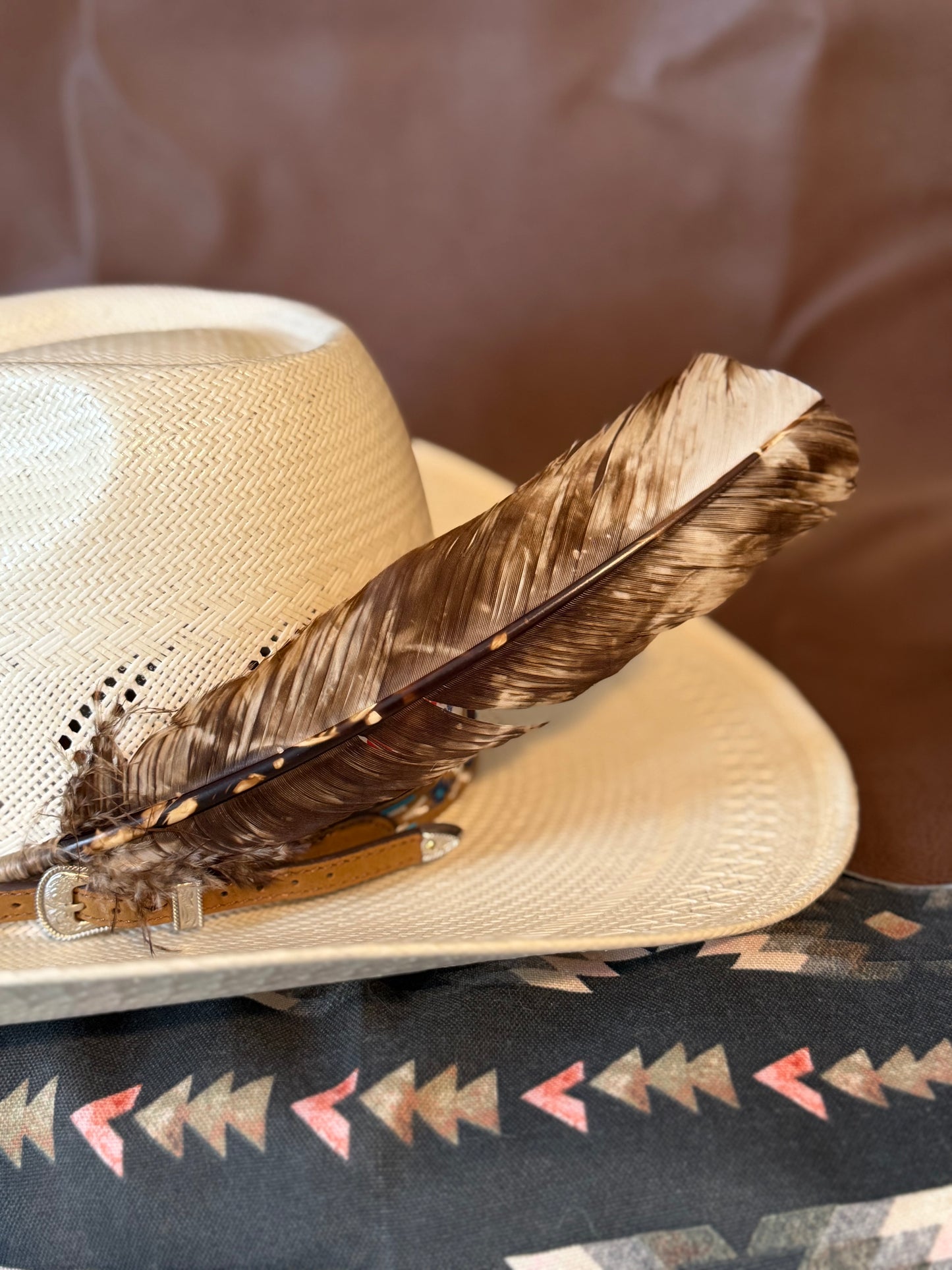A light beige woven cowboy hat with a brown feather and a tan leather strap resting on a patterned textile against a brown background.