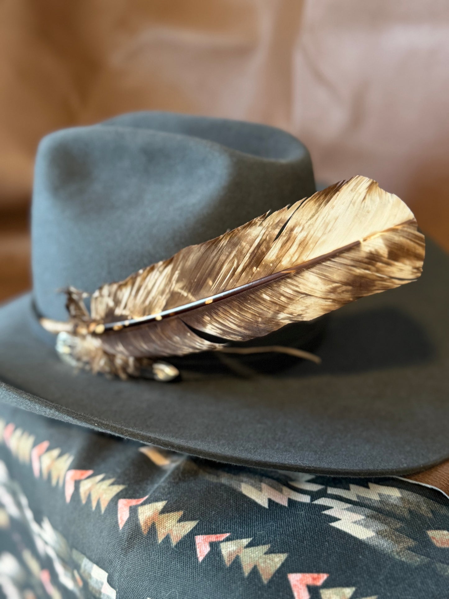 A dark gray felt cowboy hat with a large brown and beige feather resting across the brim, displayed on top of a patterned fabric surface. Large brown and cream turkey quill feather styled on a Western hat band