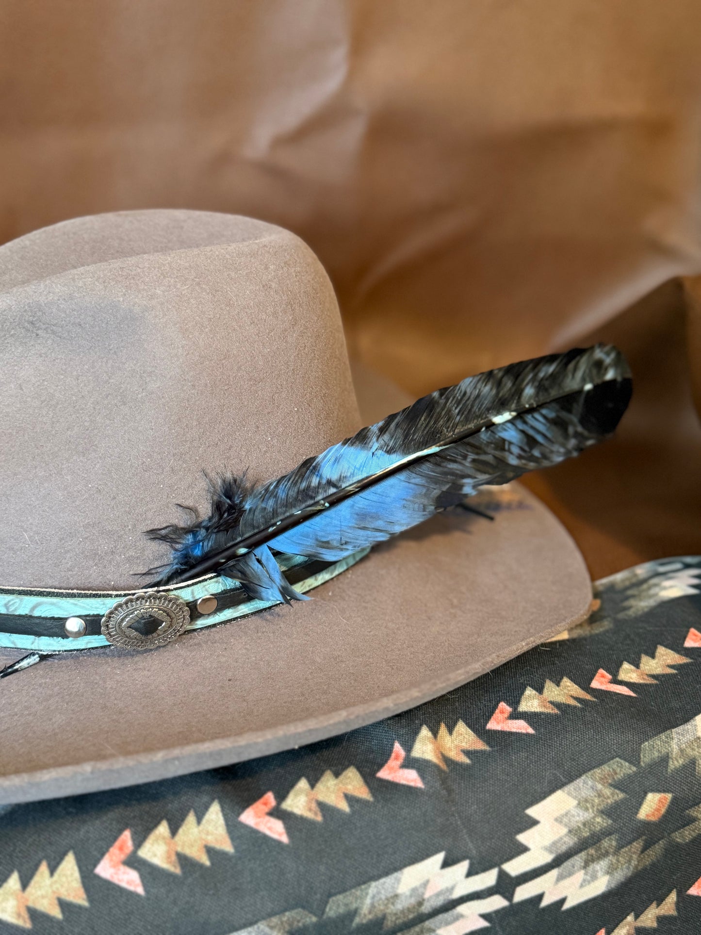 A close-up of a light brown felt cowboy hat with a turquoise and black feather band and a patterned textile underneath on a brown paper background.