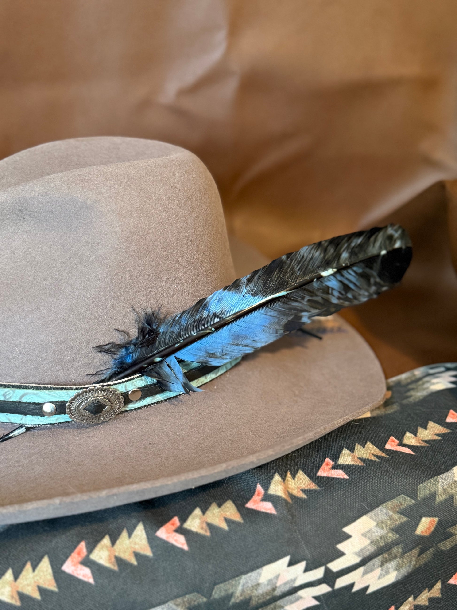 A close-up of a light brown felt cowboy hat with a turquoise and black feather band and a patterned textile underneath on a brown paper background.