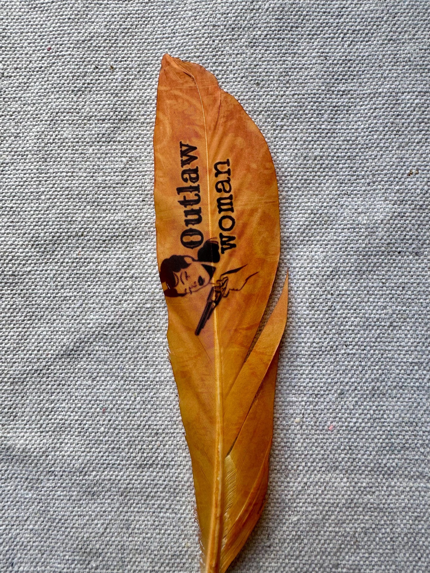 Brown feather with 'Outlaw Woman' text and graphic on a gray fabric background