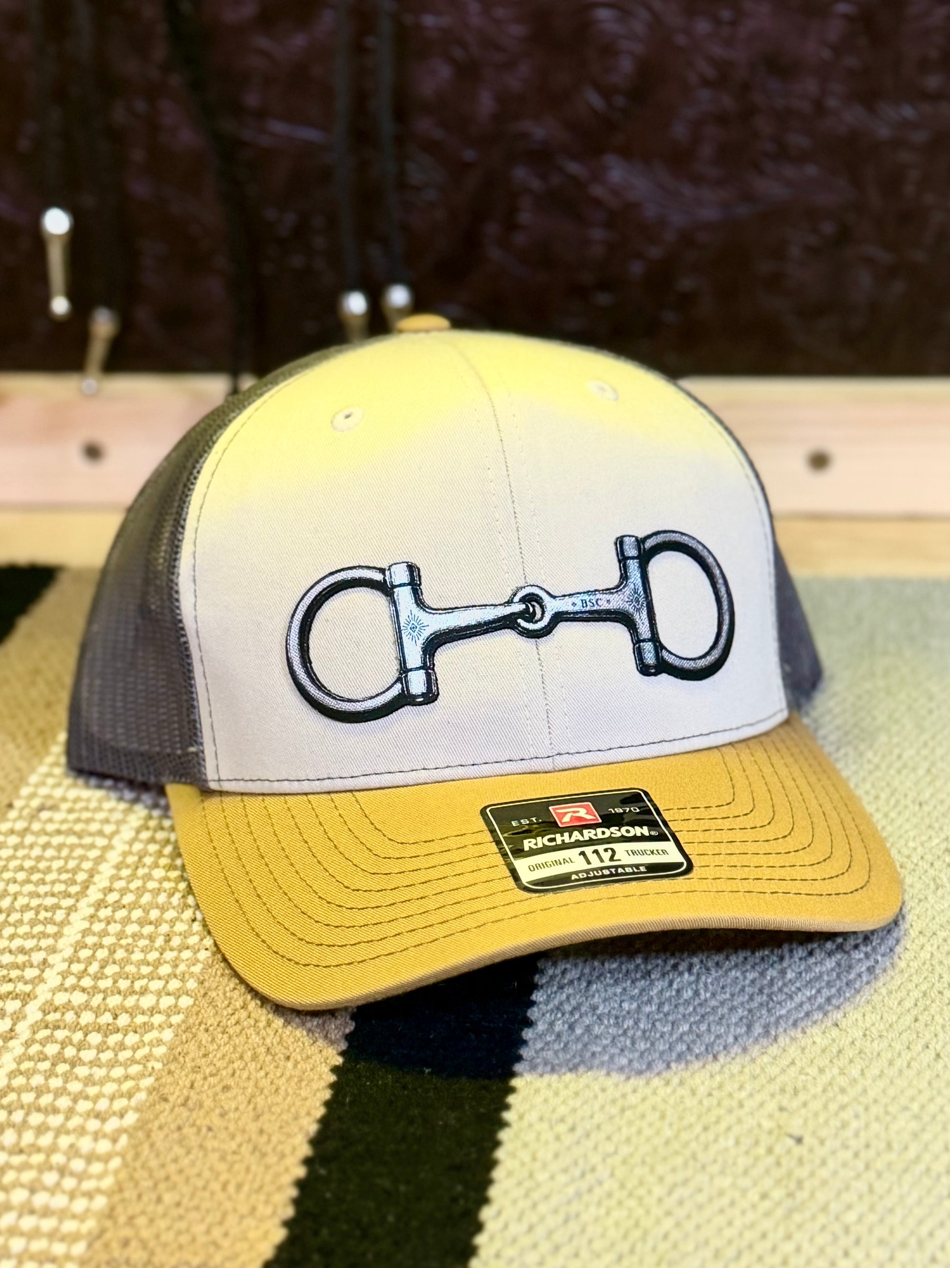 A trucker-style baseball cap with a white front panel, yellow brim, and dark gray mesh back, featuring a large black horsebit graphic on the front. A brand patch on the brim reads 'RICHARDSON.