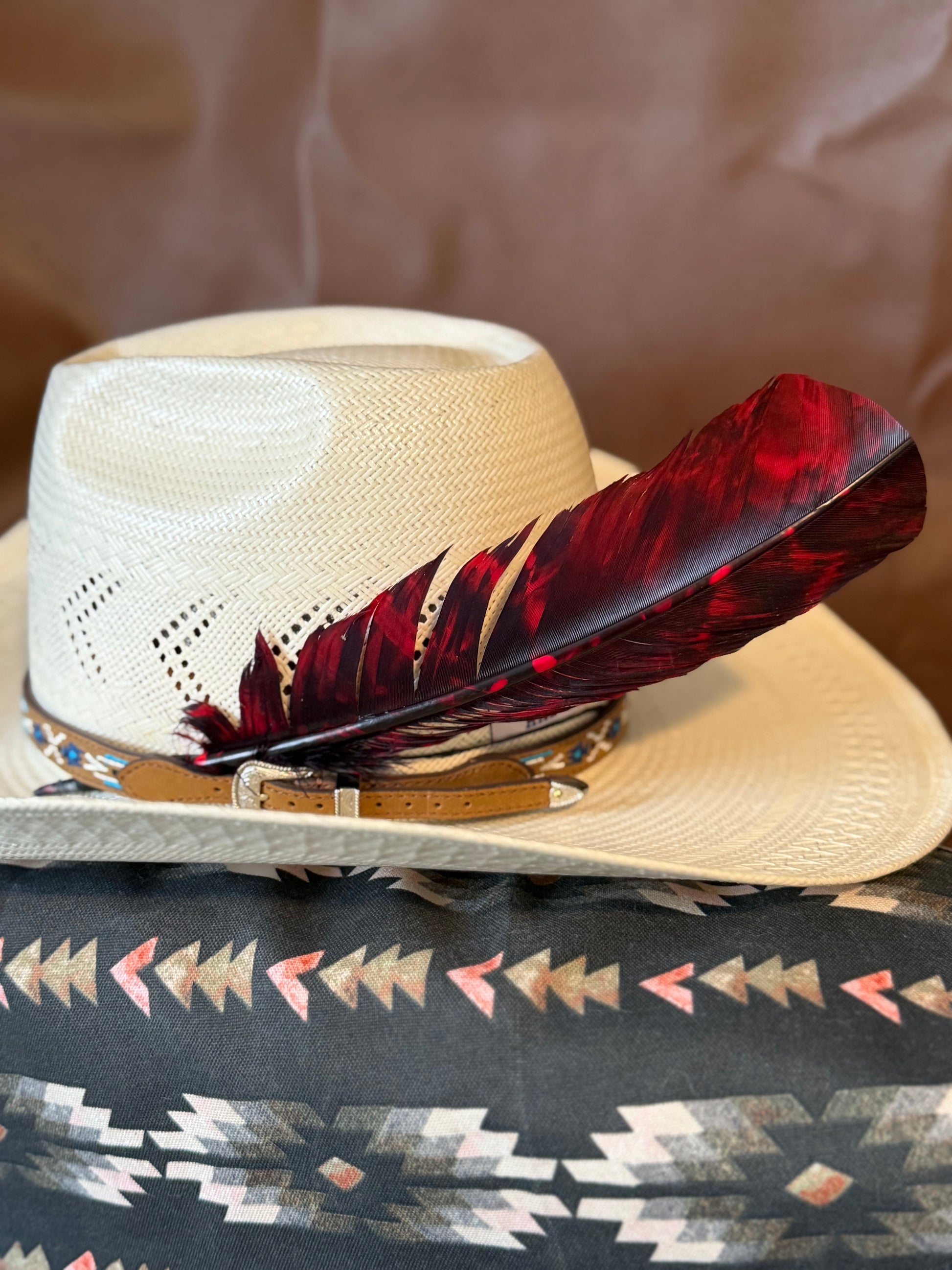 Red and black marbled Western hat feather by Buckaroo Supply Co. A cream-colored woven cowboy hat with a decorative red and black feather and a patterned band, resting on a Southwestern-style patterned blanket.