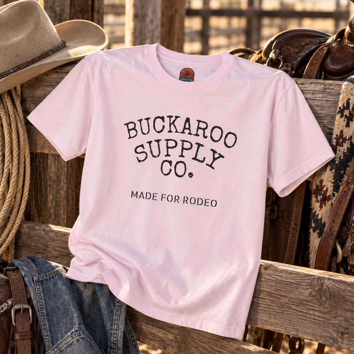 A light pink short-sleeve t-shirt with 'BUCKAROO SUPPLY CO. MADE FOR RODEO' printed in black on the front, styled on a rustic wooden bench with a cowboy hat, jeans, and western gear in a rodeo setting.