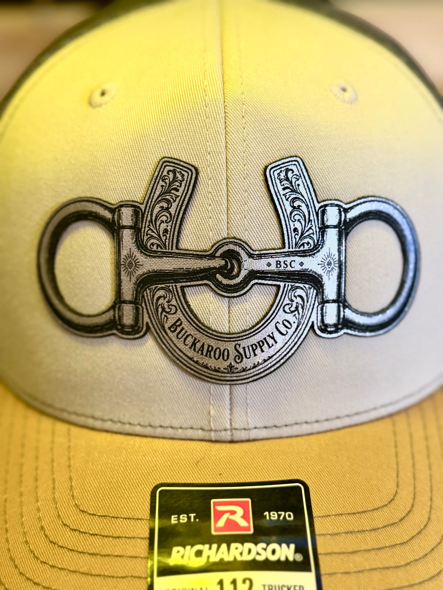 A close-up of a beige trucker cap featuring a large black and silver horse bit graphic on the front, with a yellow brim and a Richardson 112 sticker visible at the bottom.