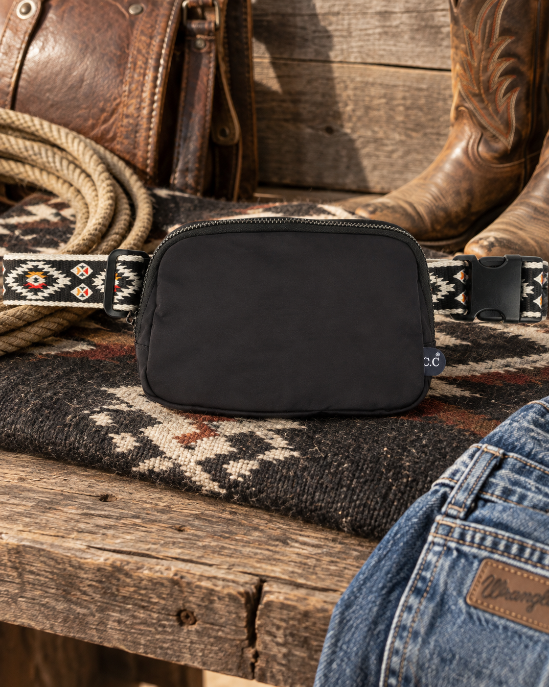 Black Southwestern strap belt bag displayed with cowboy boots, saddle, rope and denim