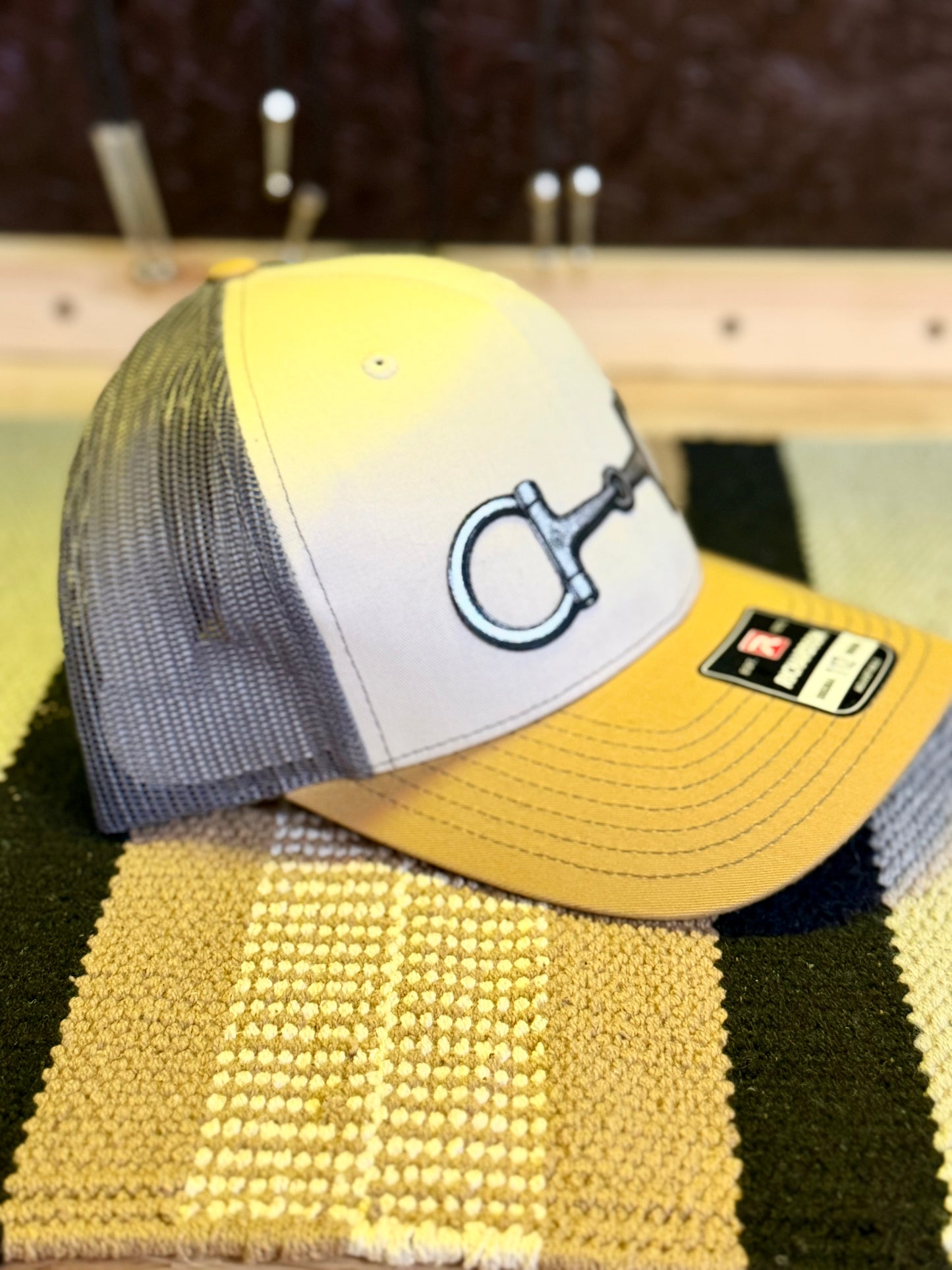 A trucker-style baseball cap with a yellow-to-white gradient front panel, gray mesh back, and a large silver horse bit logo on the front. 
