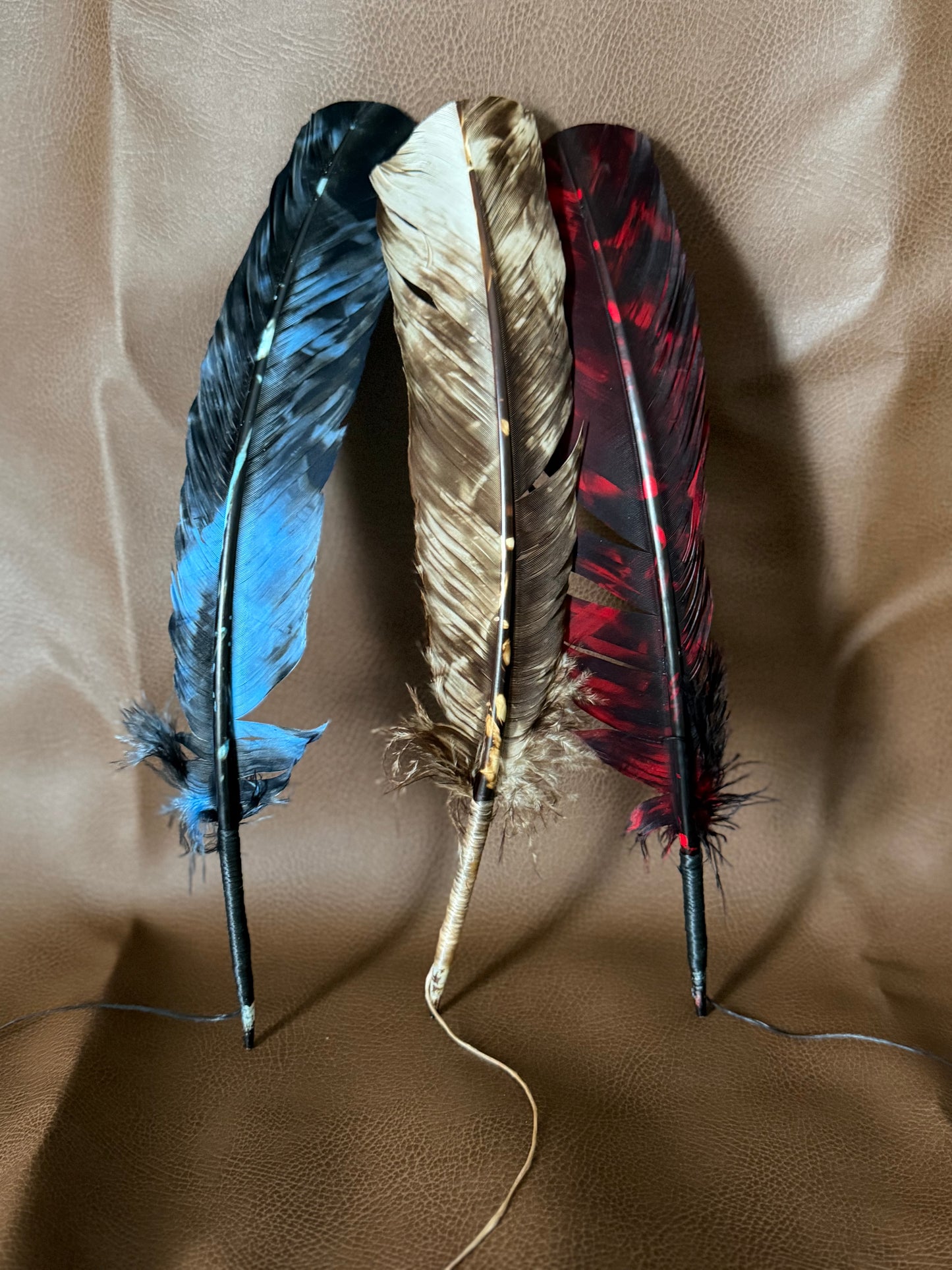 Three decorative feathers with dark shafts are arranged upright on a brown textured fabric background. The left feather is blue and black, the center feather is tan and brown, and the right feather is red and black.