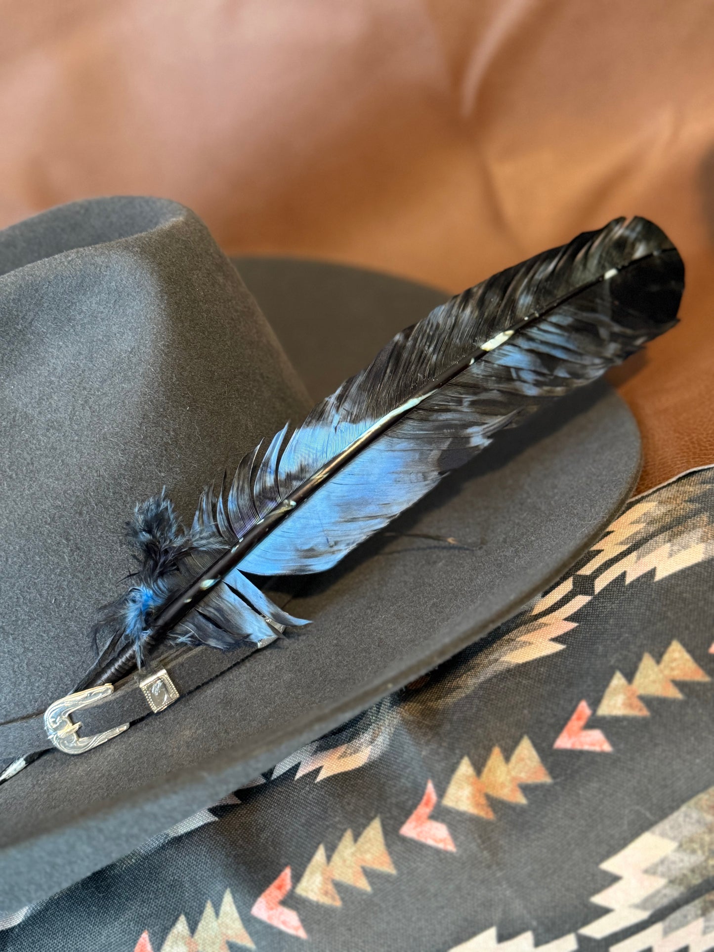 A dark gray cowboy hat with a long black and blue feather, resting on a patterned blanket with a brown leather-like background.