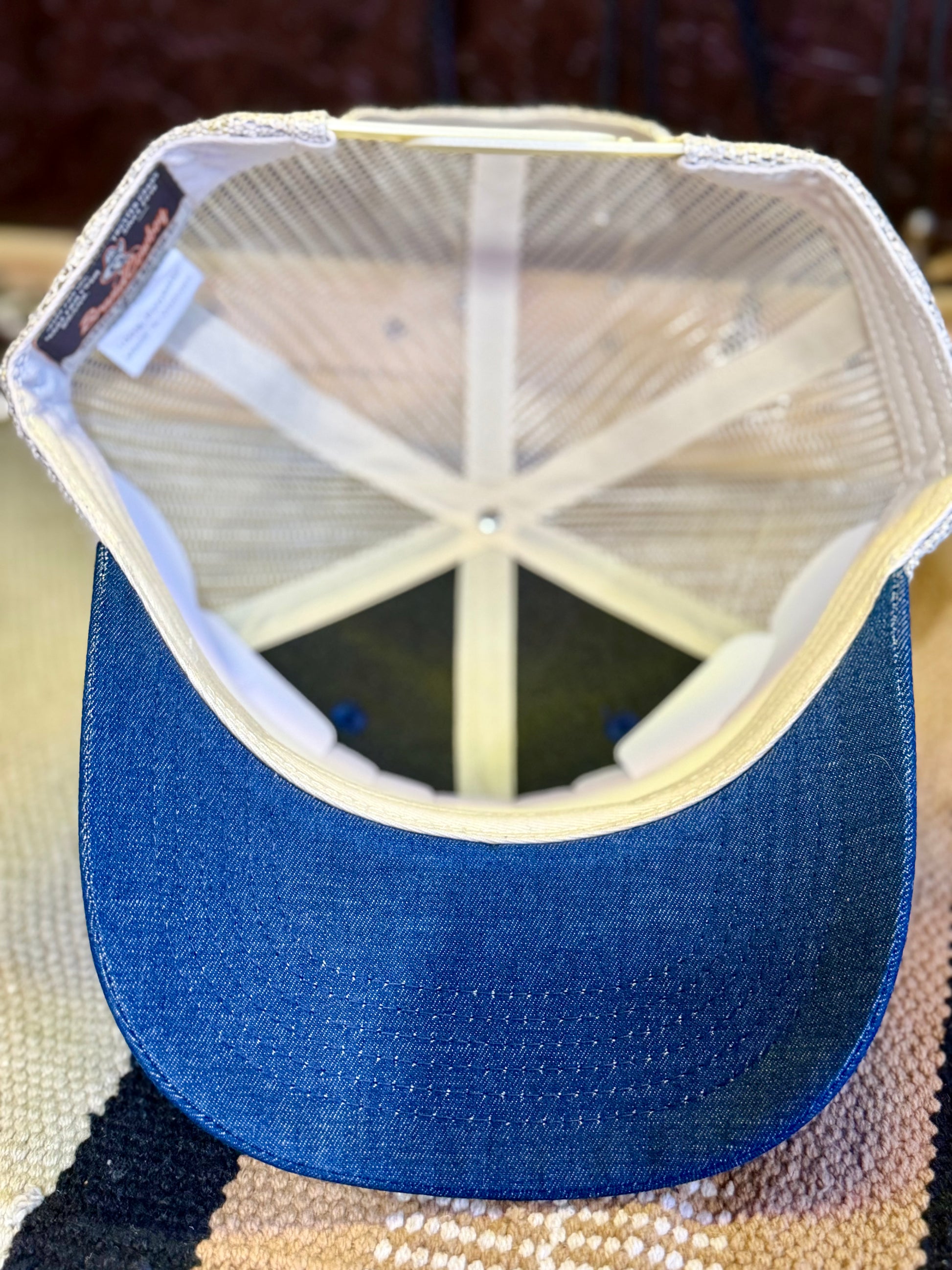 A blue denim trucker-style cap shown from the back, featuring a blue fabric brim and a cream mesh back with a white stitched edge.