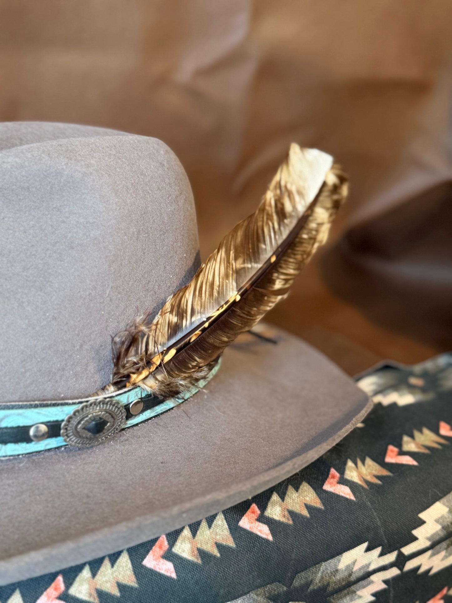 A close-up of a taupe gray felt cowboy hat featuring a brown feather with gold and black barbs and a turquoise and brown concho strap.