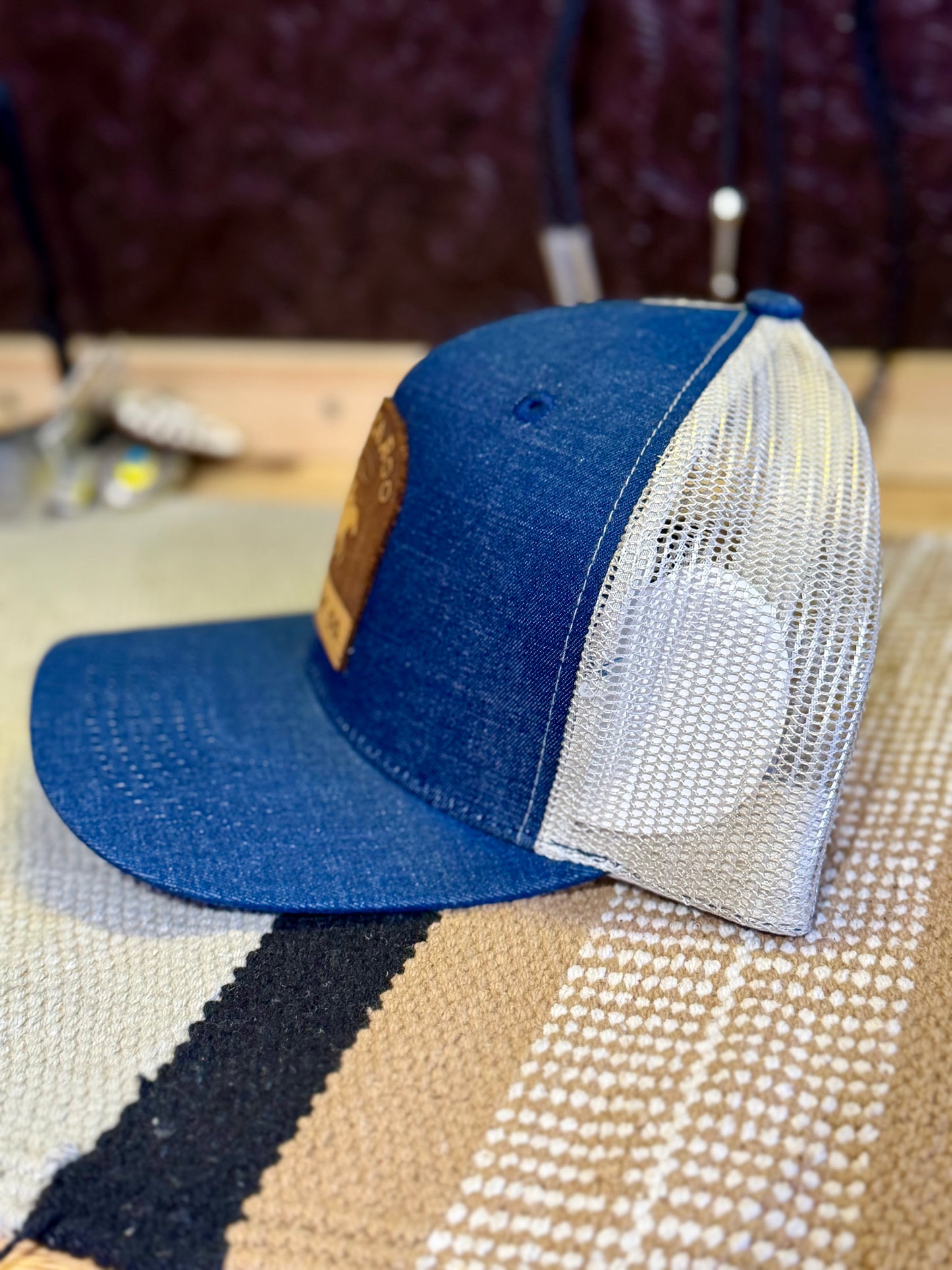 A blue denim trucker cap with a white mesh back, resting on a textured beige and black rug.