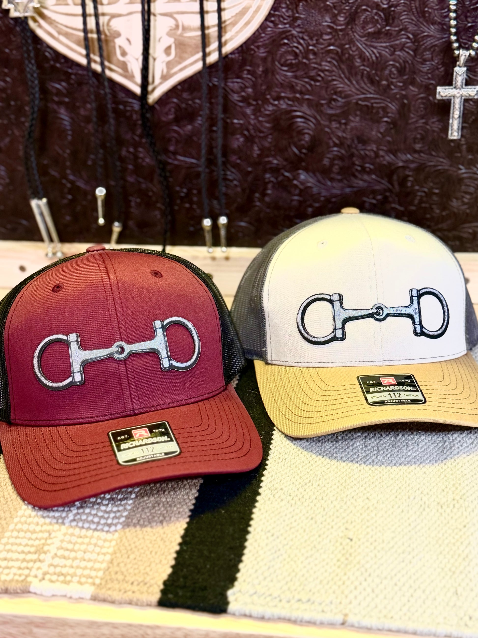 Two trucker-style caps with horsebit patch on the front panels, displayed side by side on a textured surface. The left cap has a cardinal red brim and black mesh back, while the right cap has a cream brim, black mesh back, and mustard yellow visor.