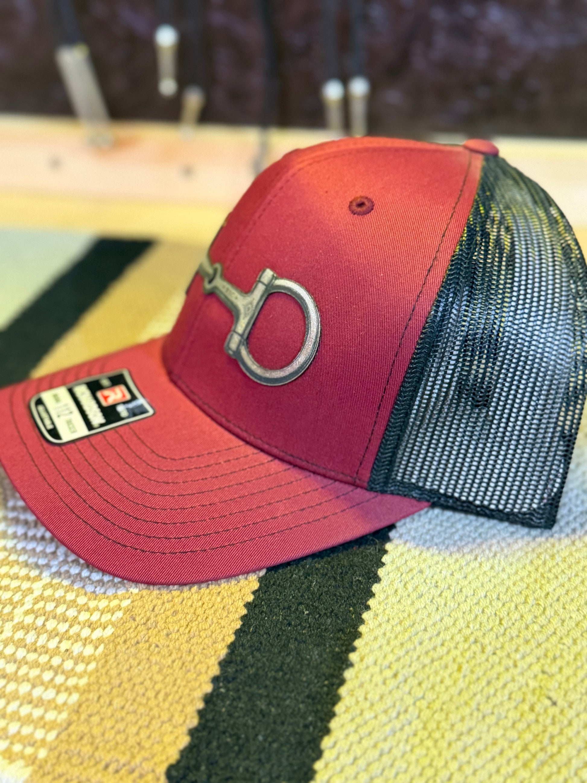 A red trucker-style baseball cap with a black mesh back and a silver metal horsebit logo on the front, resting on a yellow and black striped woven blanket.