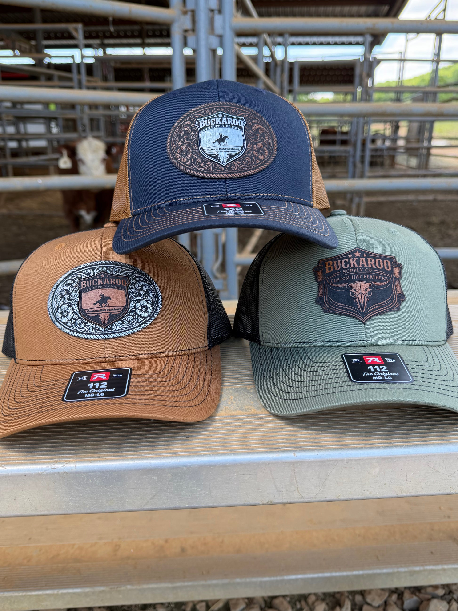 Three caps in blue, green, and brown with logos on a wooden surface. Loden (sage) Green and black, Richardson 112 cap with Buckaroo Supply Co logo.