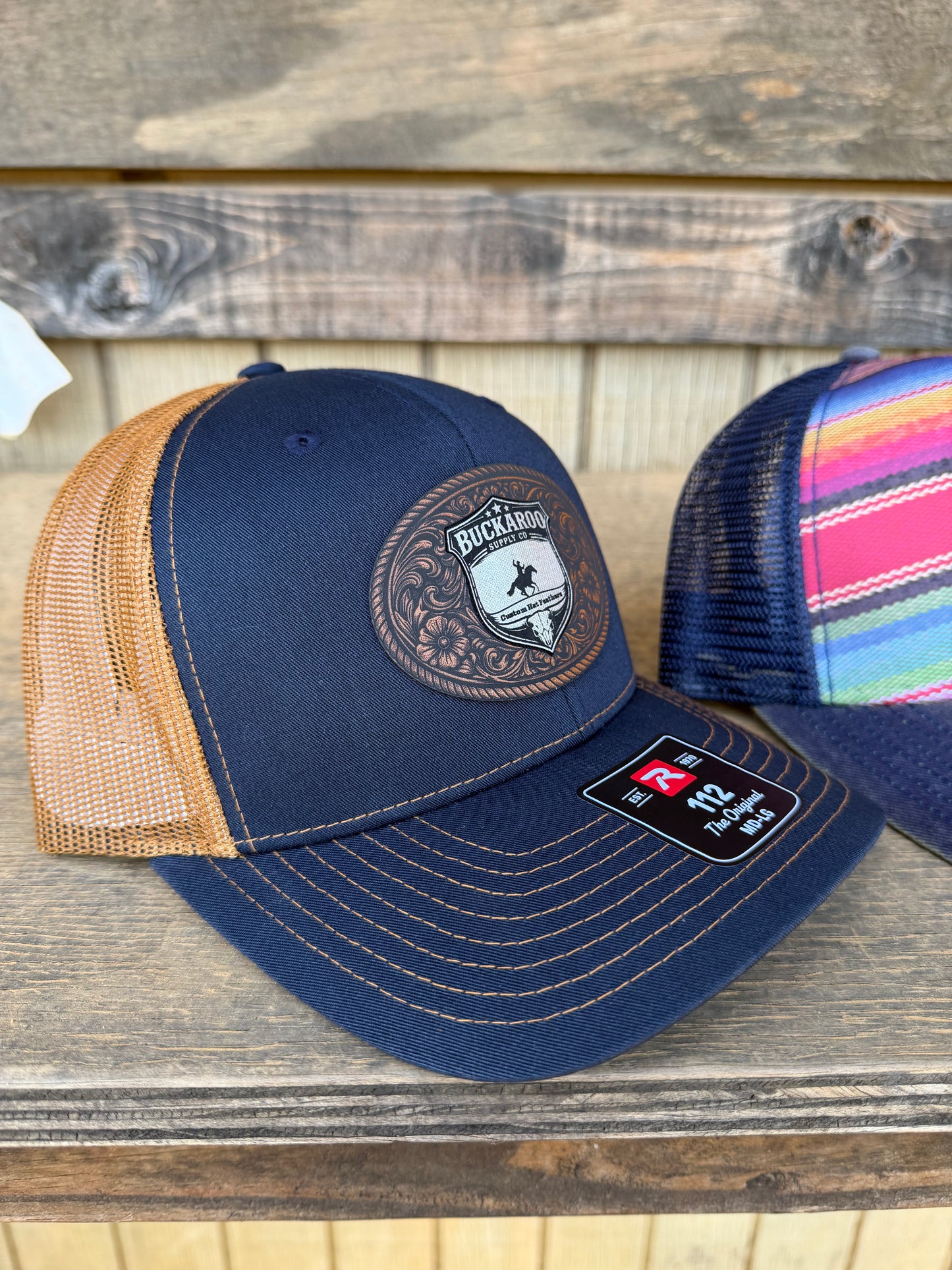 Navy blue cap with brown mesh and colorful strap on a wooden surface. Navy and Camel Richardson 112 Western Trucker Hat with Buckaroo Supply Co. logo.