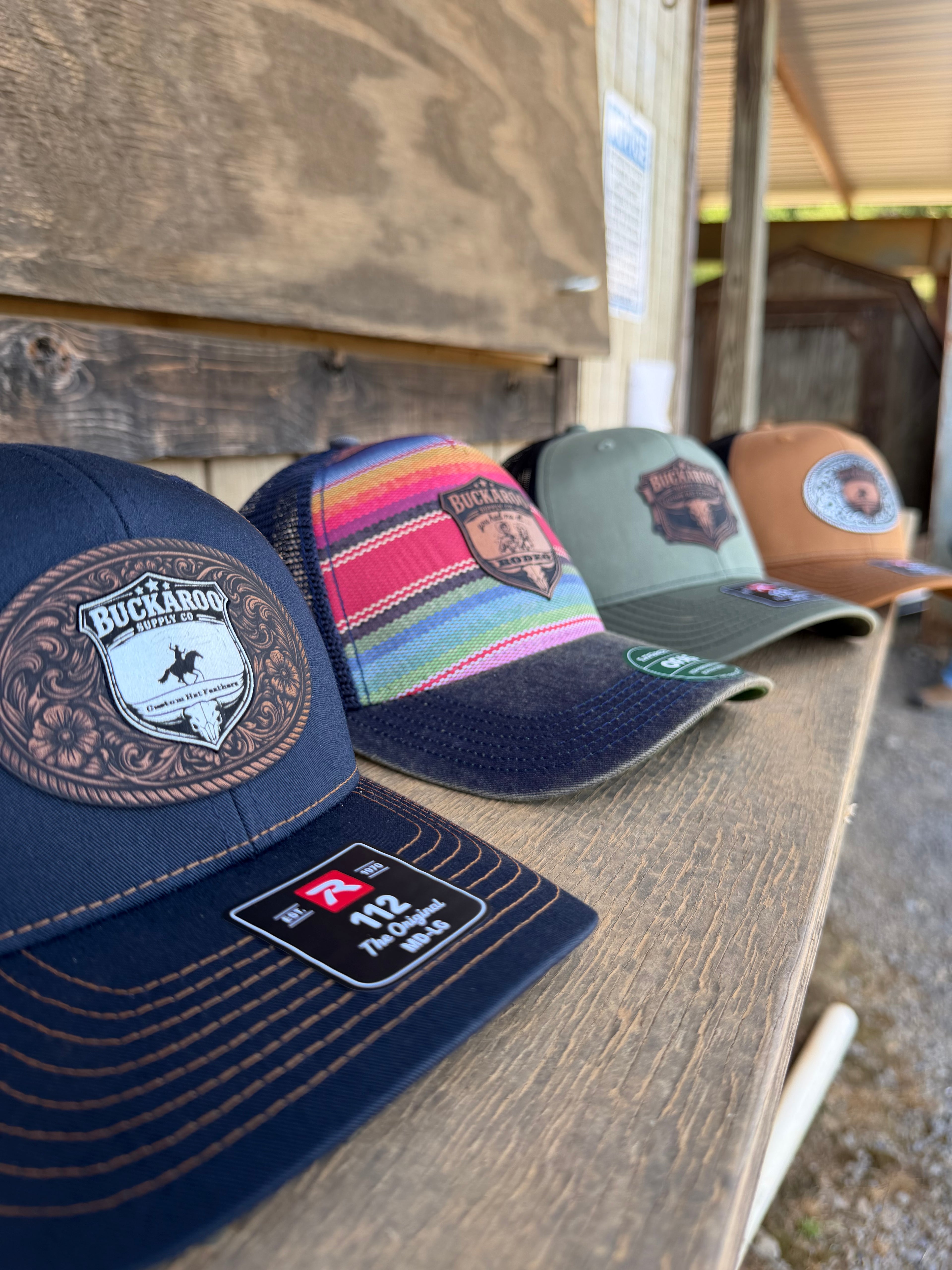 Four baseball caps with different designs and colors on a wooden surface. Richardson 112 cap with Buckaroo Supply Co logo.