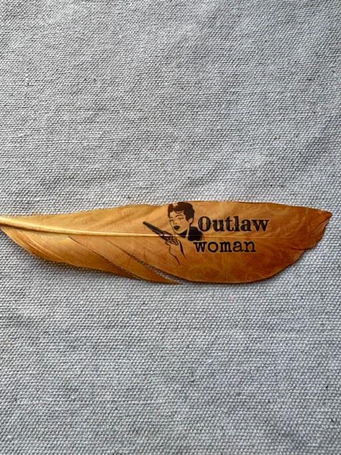 Brown feather with 'Outlaw woman' engraving on a gray fabric background