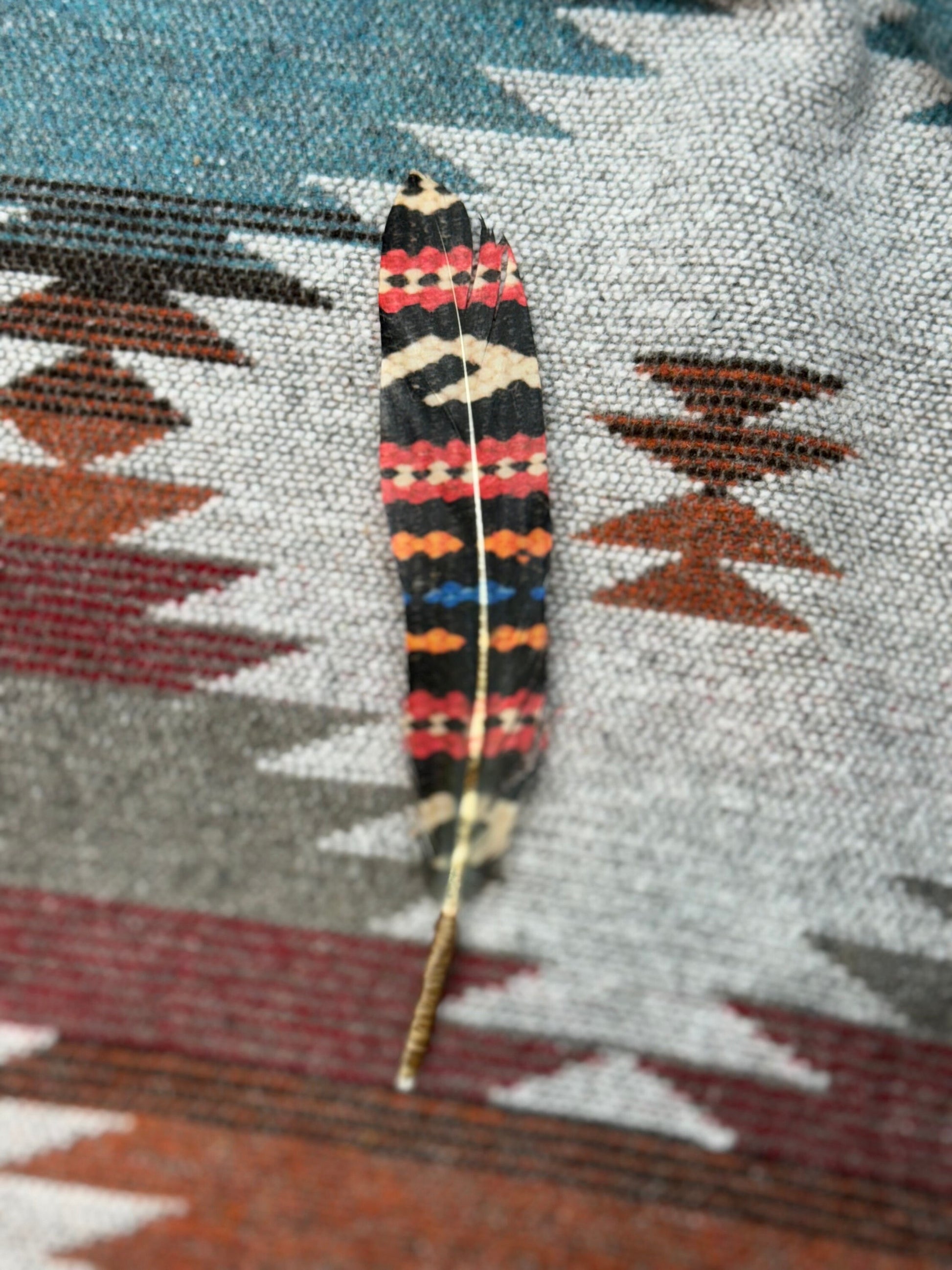 A single decorative feather with a black base and red, orange, beige, and blue geometric bands, laid on a patterned woven textile.