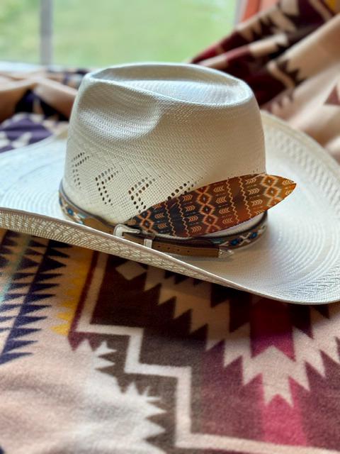 A white woven cowboy hat with a patterned brown and orange band and silver concho, resting on a southwestern patterned blanket.
