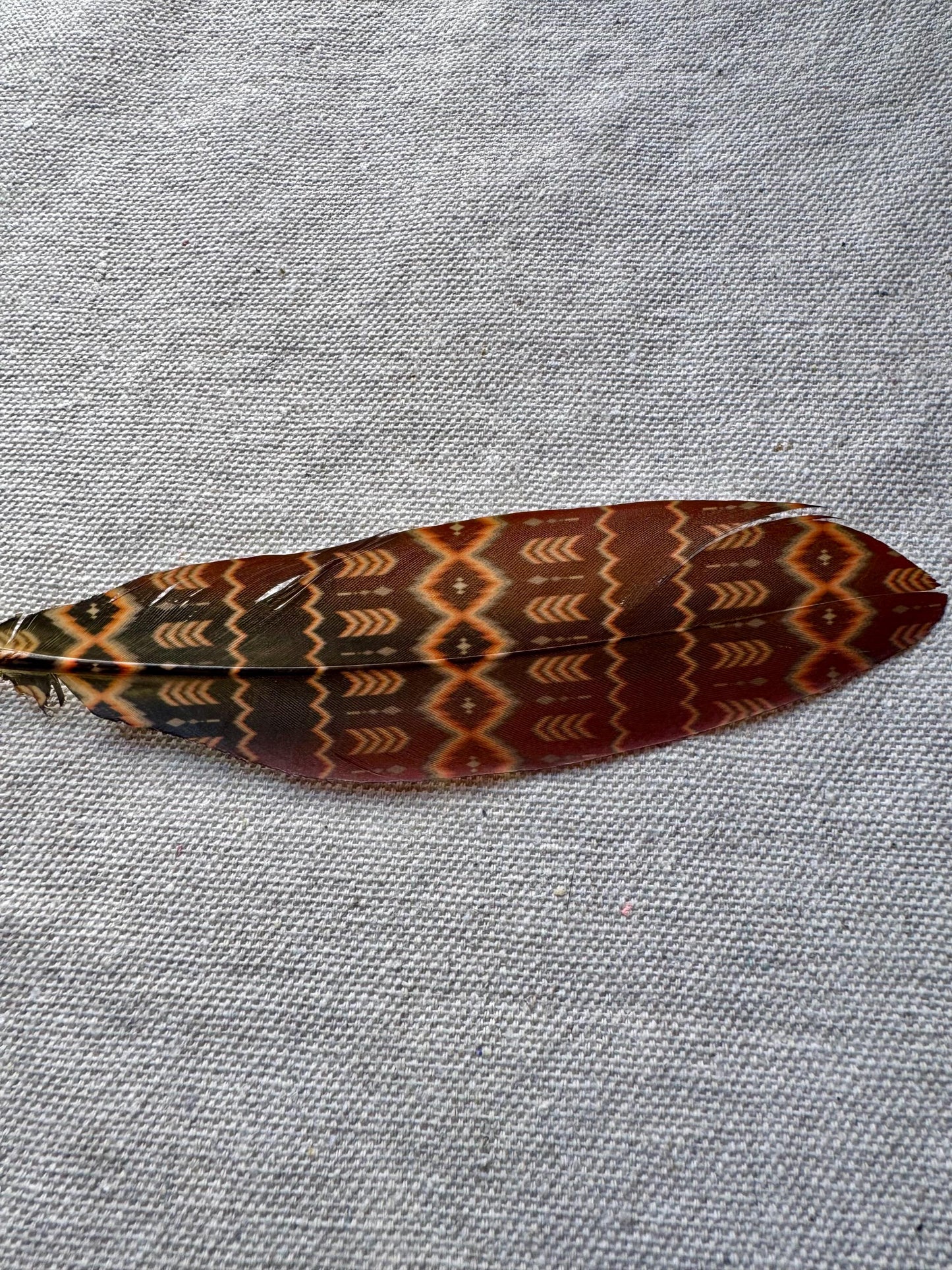 A single decorative feather with a brown, orange, and black geometric pattern, laid flat on a textured gray fabric background.