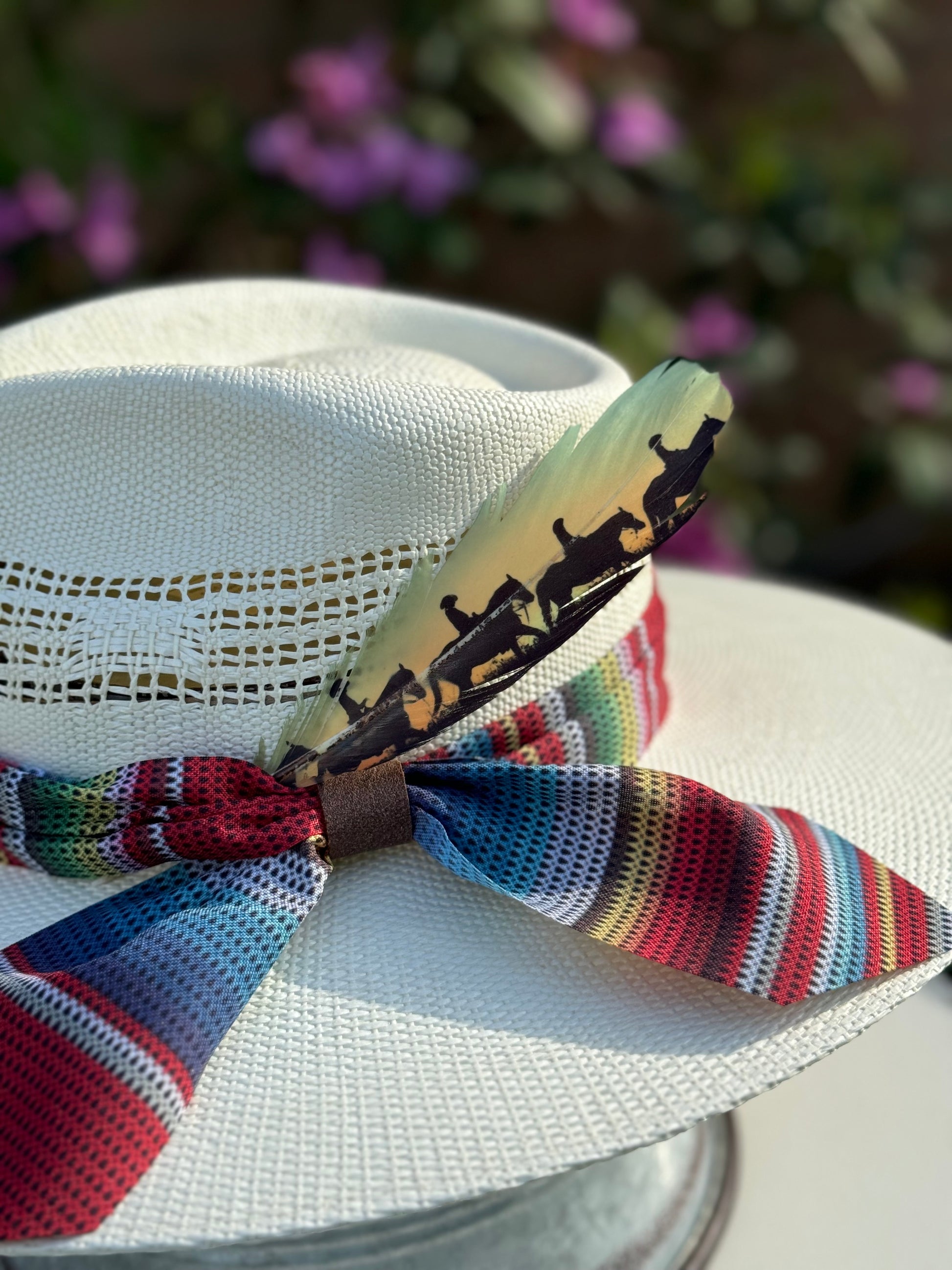 White straw hat with colorful ribbon, Buckaroo Supply Co. feather, and decorative band featuring silhouettes against a blurred floral background